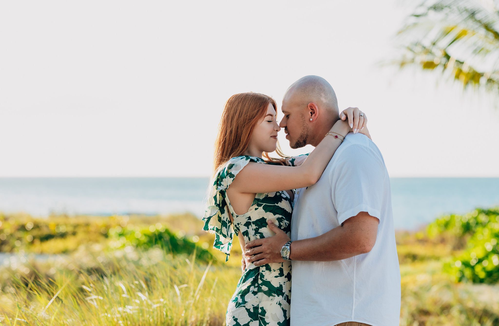 A couple is embracing tenderly on a beach with ocean in the background during sunset, both with eyes closed, foreheads touching, in a romantic moment.