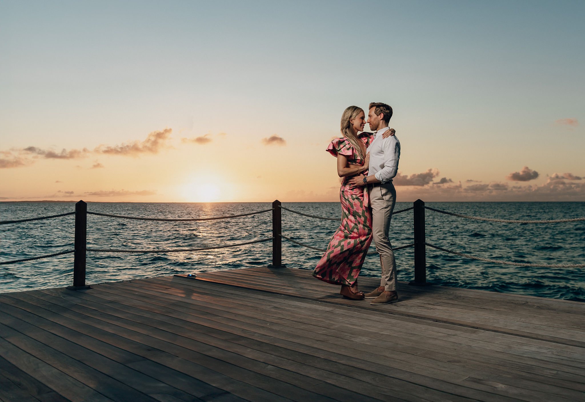 A couple stands close together on a wooden dock at sunset, overlooking the ocean, embracing and smiling at each other.