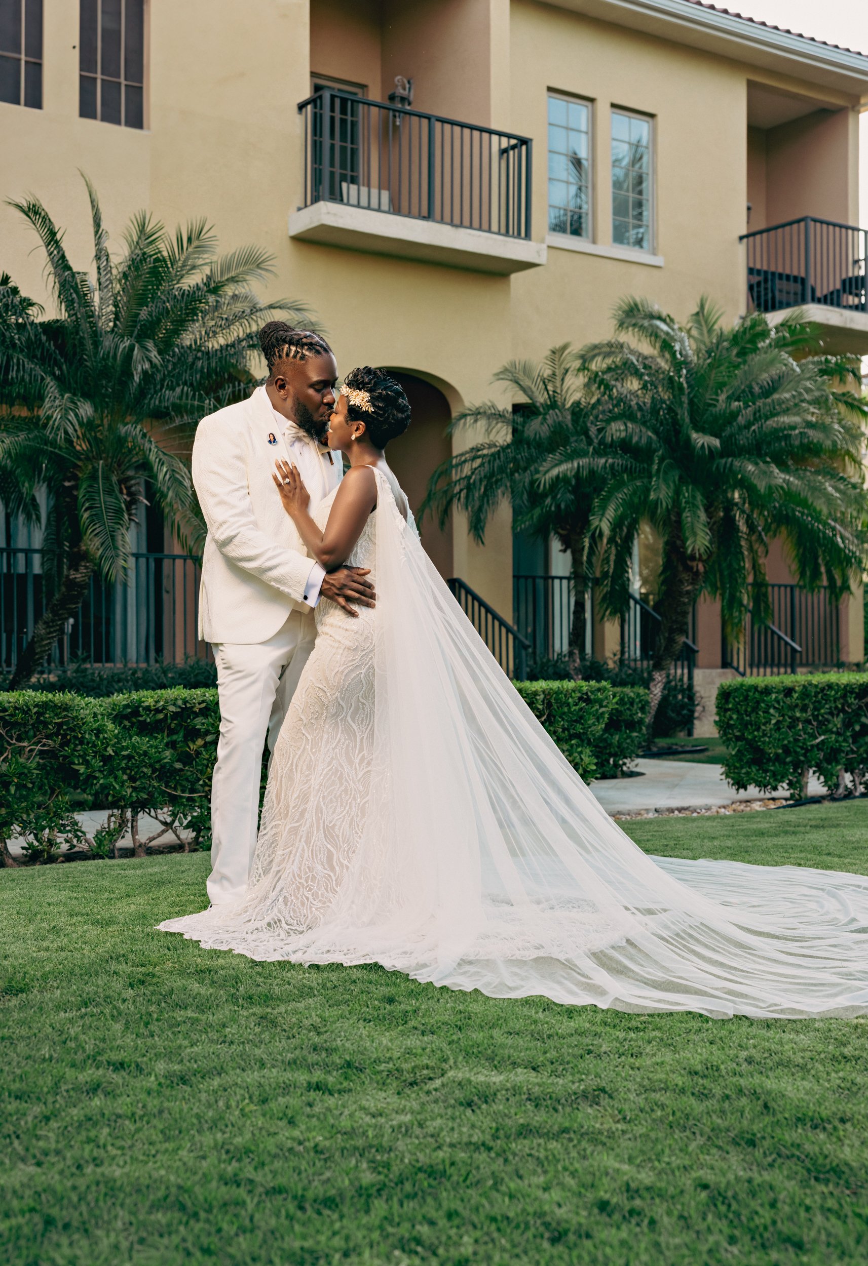 Couple dressed in wedding attire sharing a kiss outdoors in front of a yellow building with palm trees.