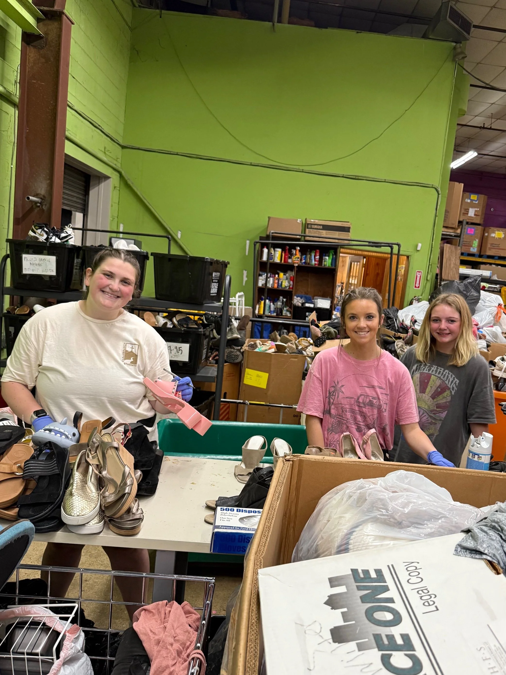 Three smiling young women working at a table with shoes and boxes in a warehouse or thrift store. The background has shelves filled with various items, and the wall is painted bright green.