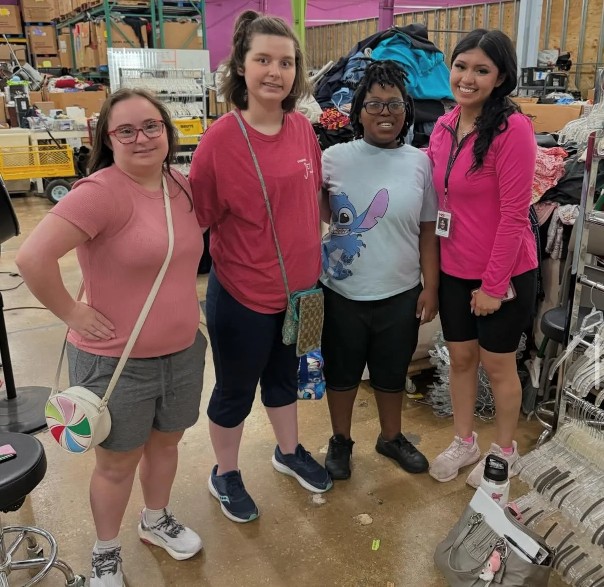 Four women standing together inside a thrift store or warehouse, surrounded by shelves filled with clothing, bags, and miscellaneous items, smiling at the camera.