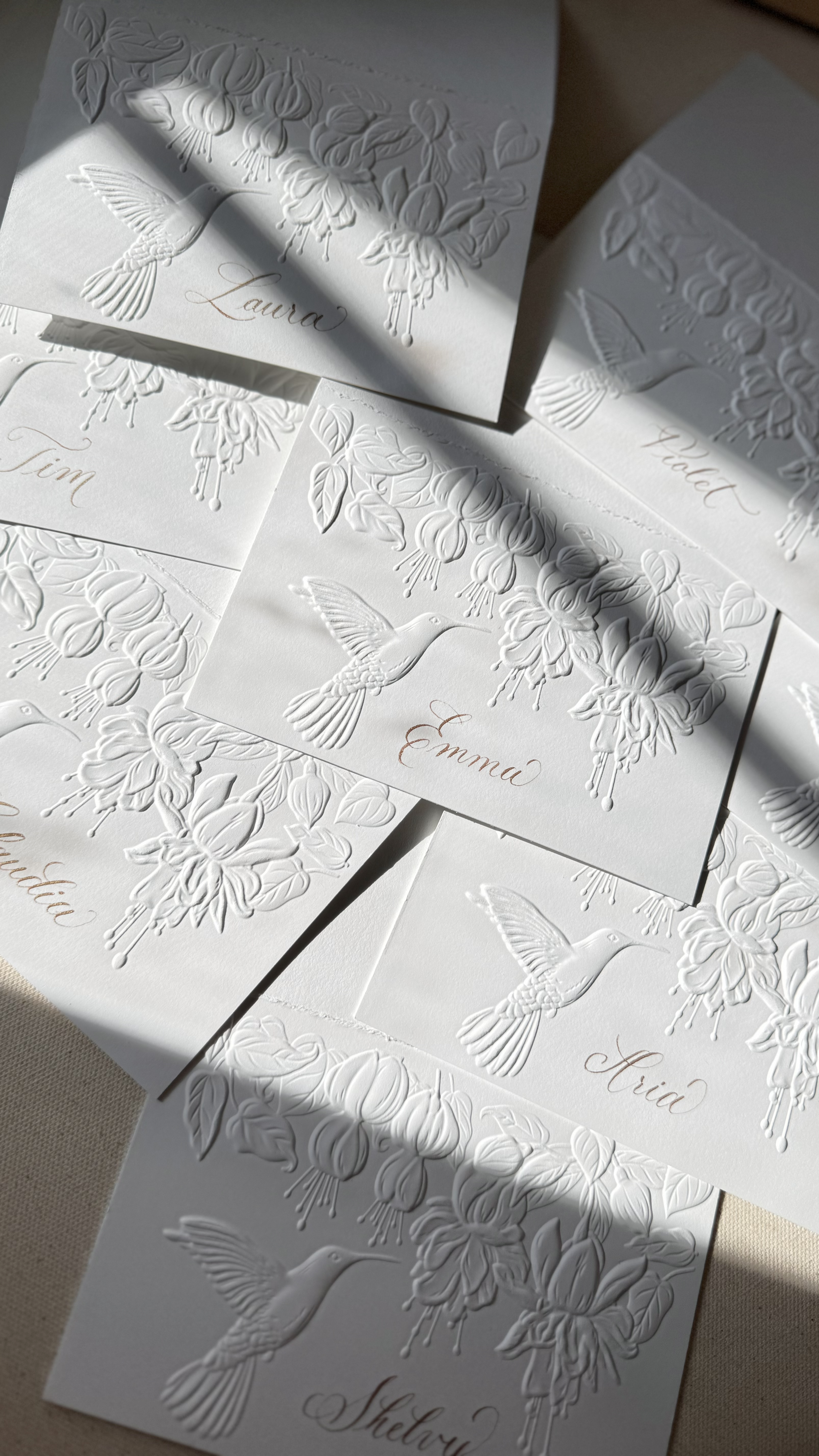 Elegant white embossed place cards with hummingbird and floral designs, featuring handwritten names in gold ink.