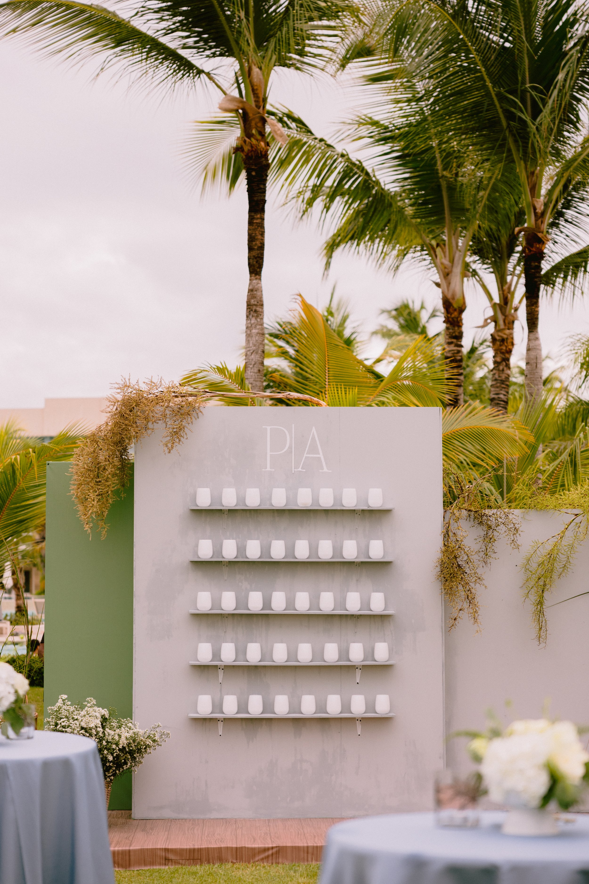 A white wall with the name 'PIA' at the top, several shelves with white cups, tropical trees in the background, and tables with floral arrangements in the foreground.