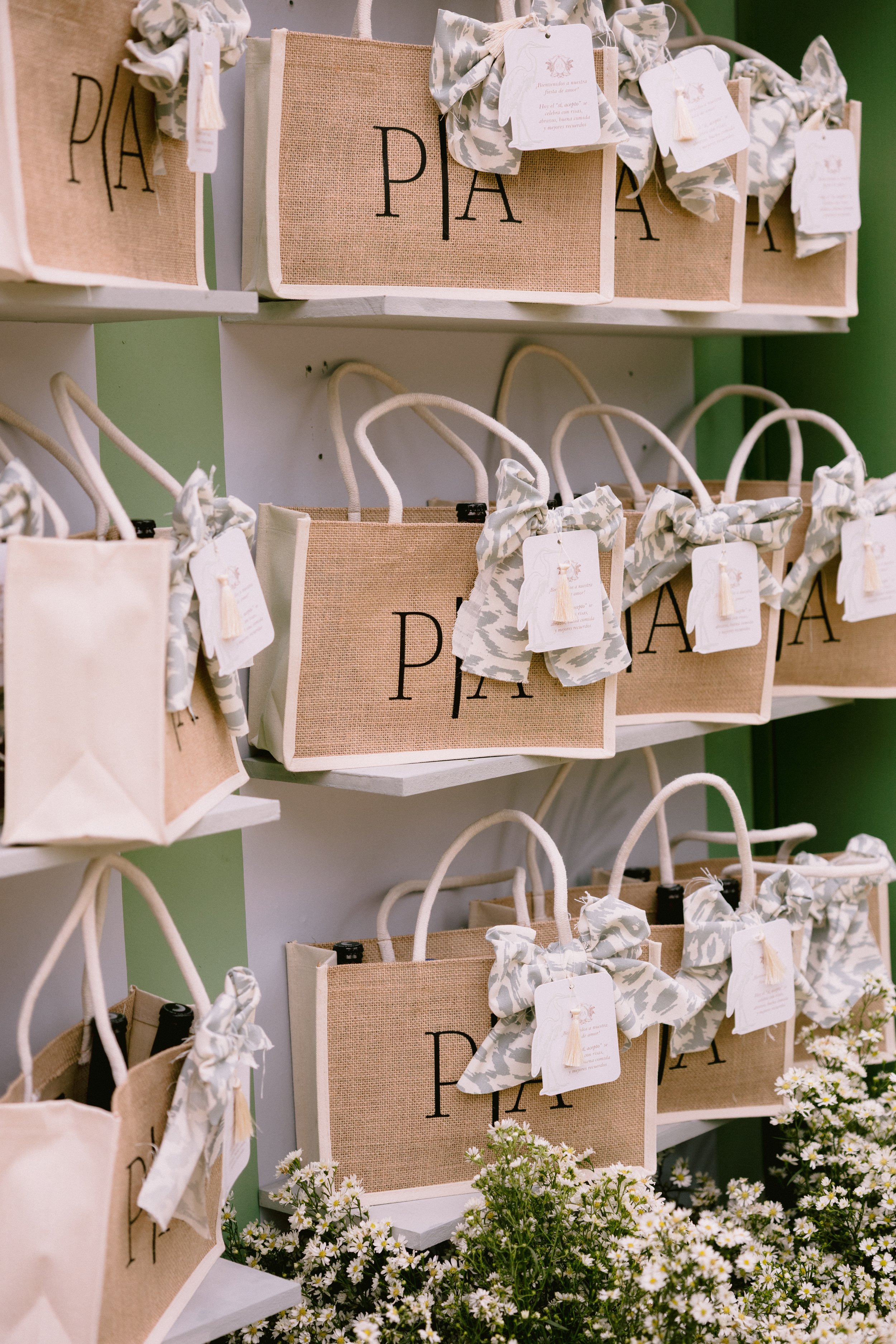 Multiple beige and white tote bags with bows hanging on white shelves, with white and yellow flowers at the bottom.