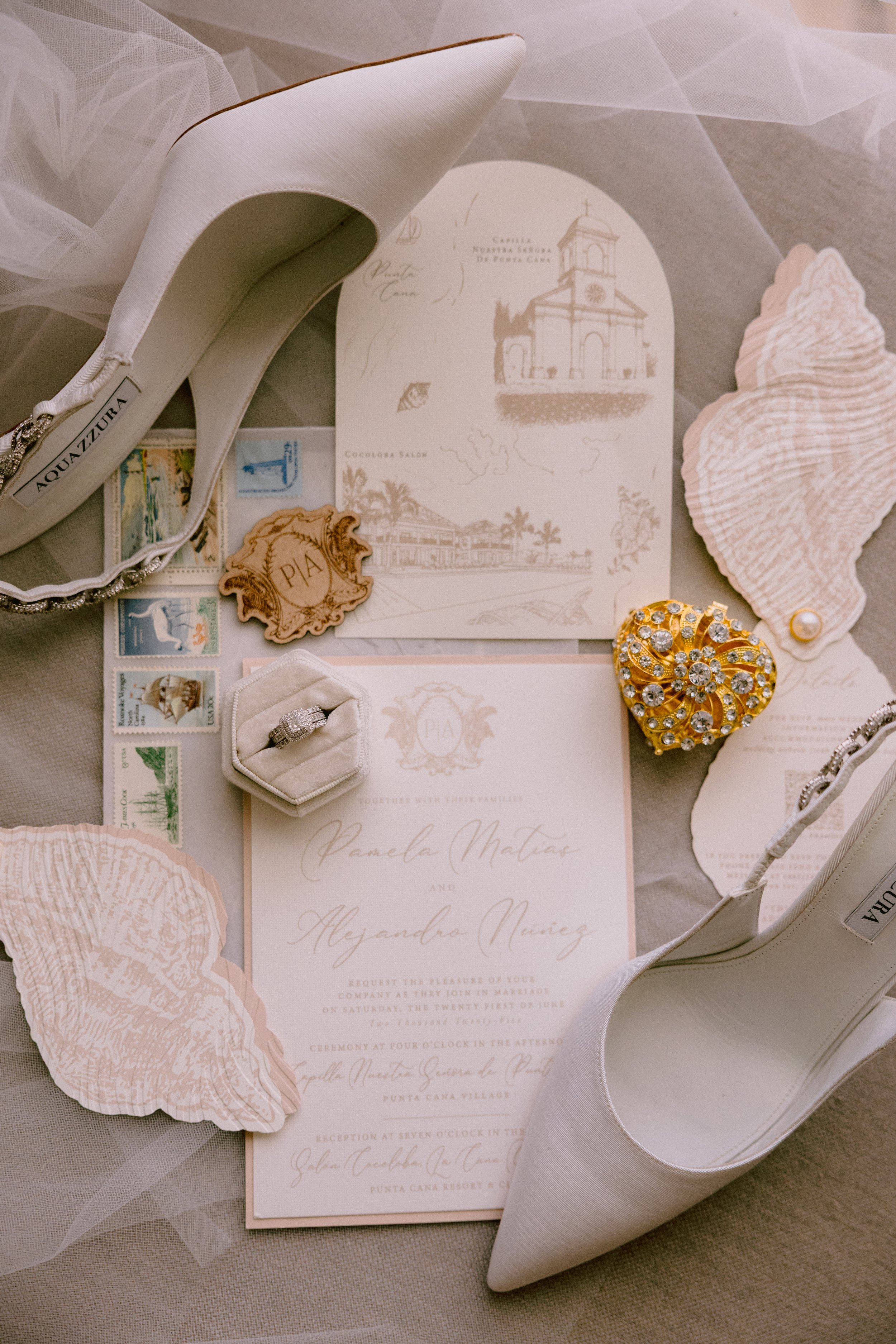 Wedding invitation surrounded by white heels, floral paper cutouts, a diamond ring, a gold decorative item, and various small aesthetic decorations.