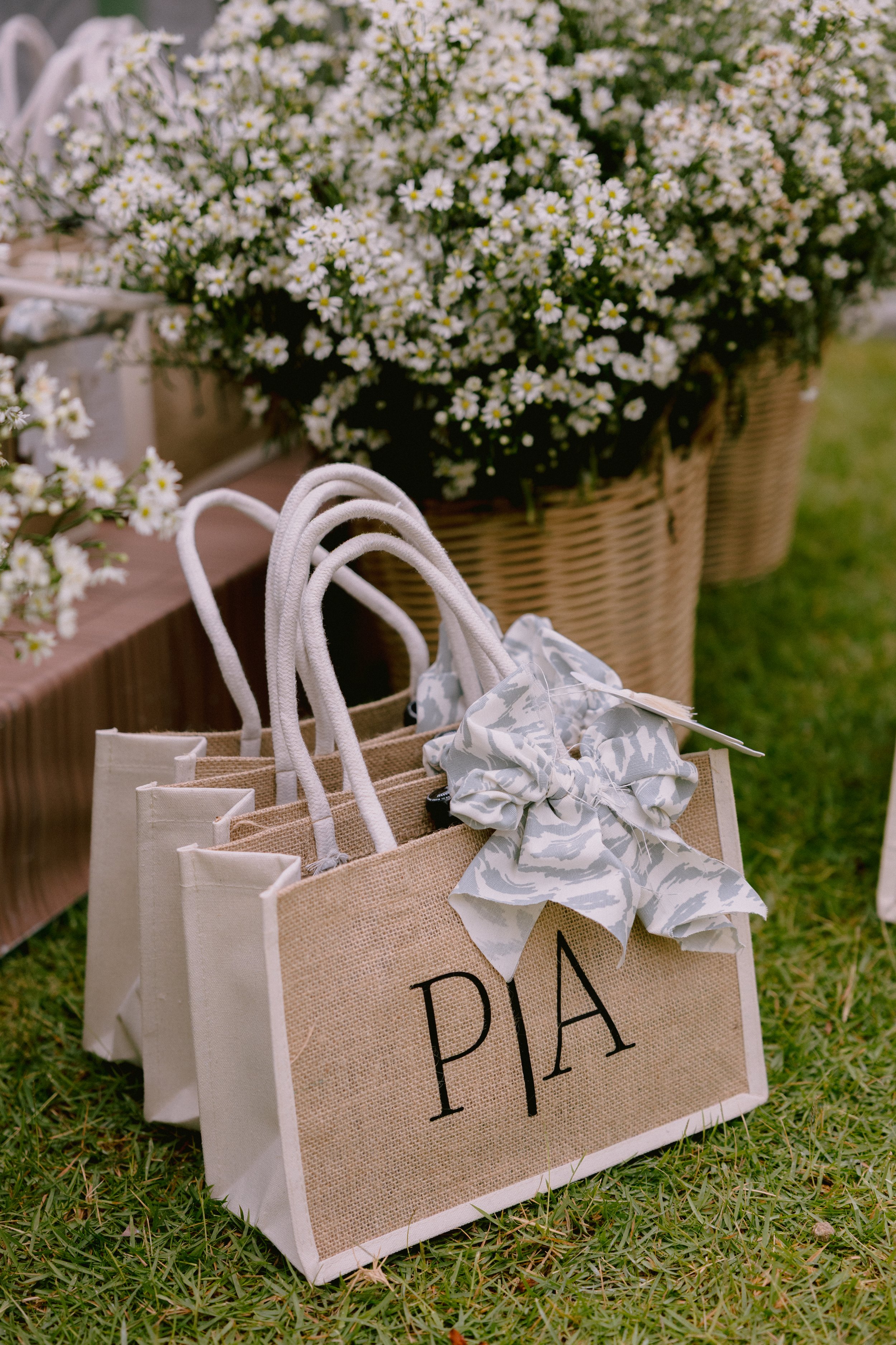 Gift bags with 'PIA' written on one bag, decorated with a gray and white bow, placed on grass with a basket of white flowers in the background.