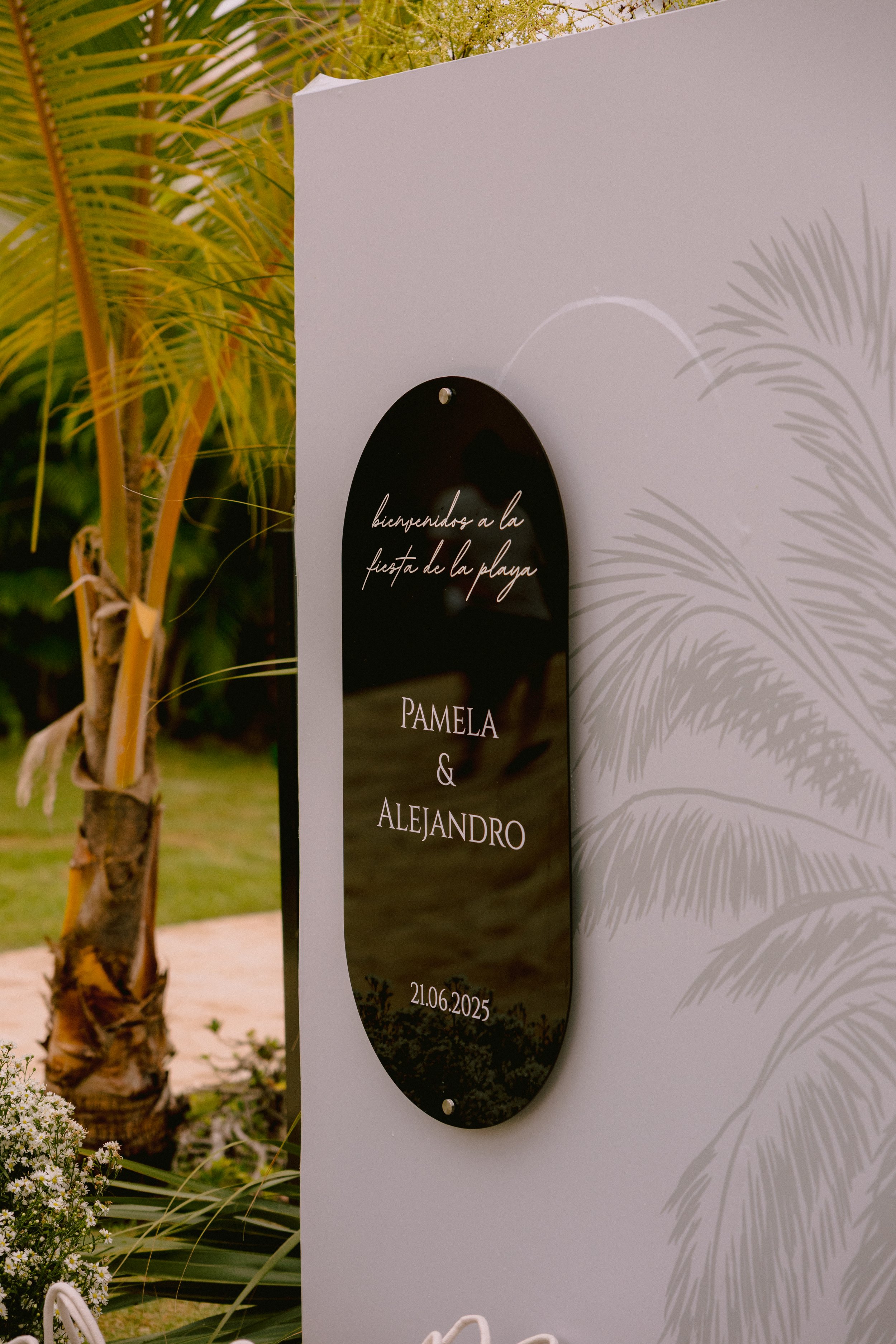 Reception sign with black rounded rectangular plaque that says 'bienvenidos a la fiesta de la playa, Pamela & Alejandro, 21.06.2025', next to white panel with palm leaf shadow design, outdoor setting with palm tree and greenery.