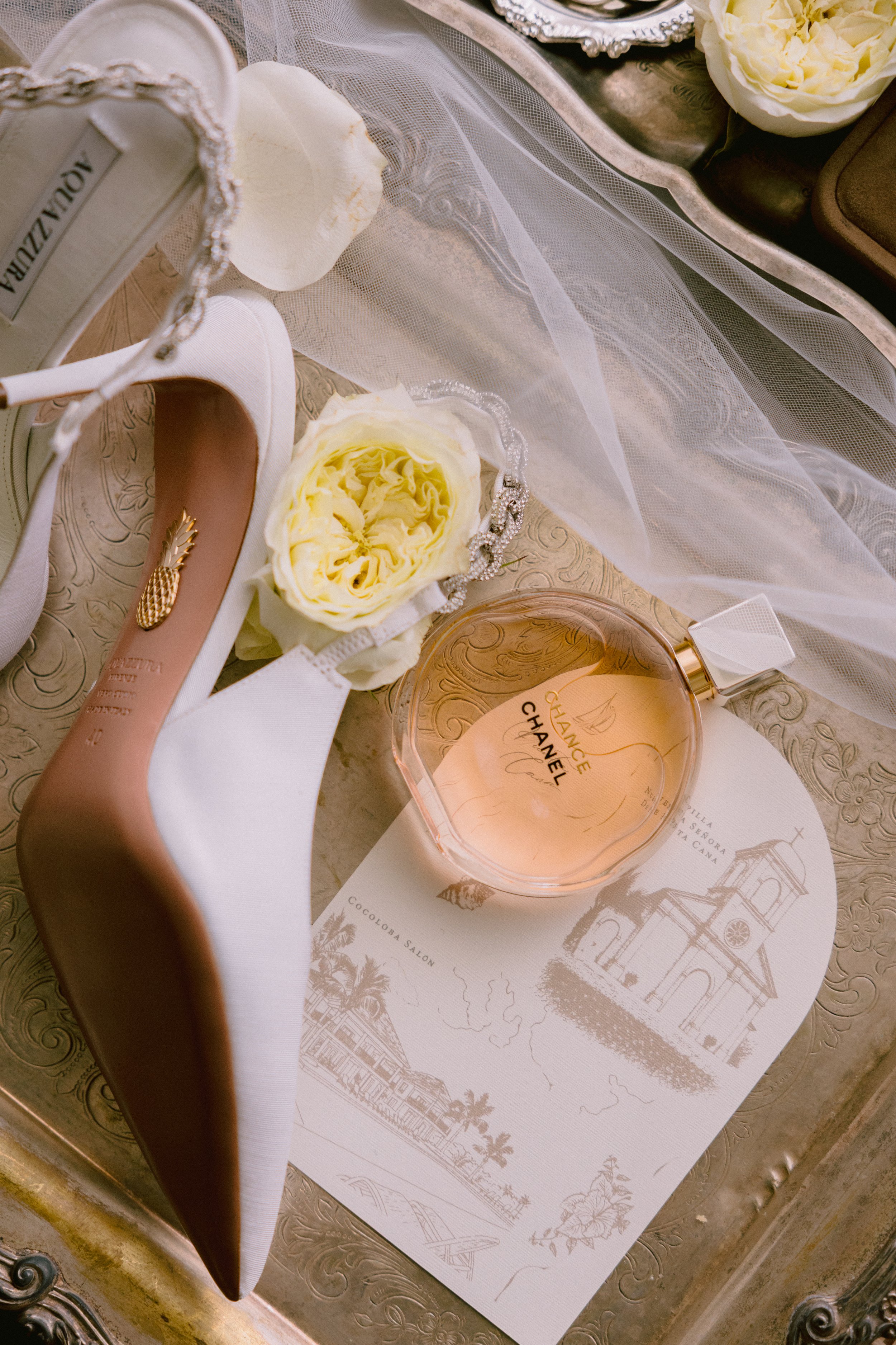 A flat lay of a white high-heeled shoe with a gold pineapple embellishment, a bouquet of white roses, a bottle of Chanel perfume, a decorative tray, and a printed paper with church and landscape illustrations.
