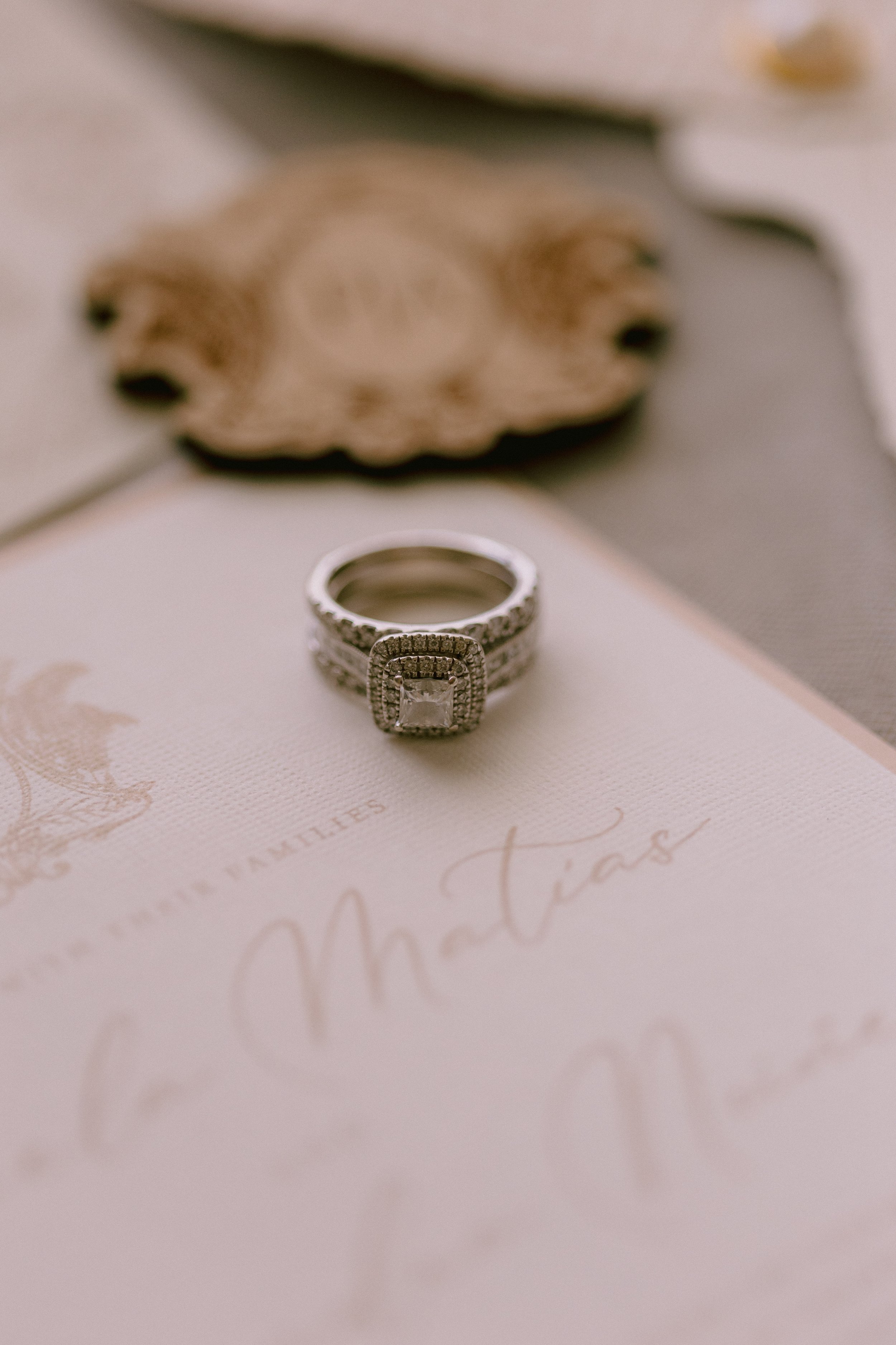 A diamond engagement ring with a square center stone and intricate band, placed on a white RSVP card with gold cursive lettering that says "Matias" and other text partially visible, with a blurred decorative envelope in the background.