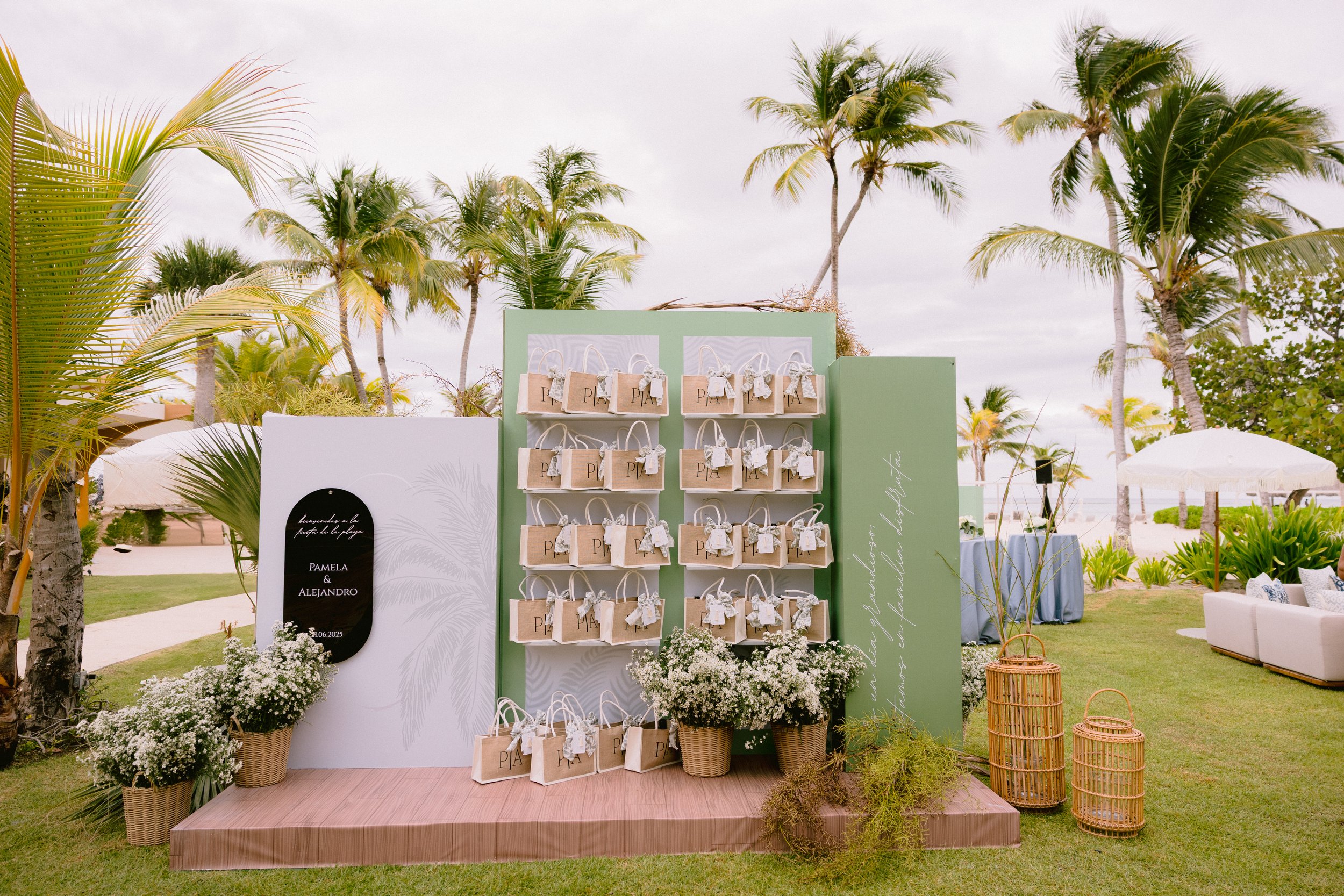 Wedding favor display with gift bags on a green and white stand, decorated with white flowers, set outdoors among palm trees under a cloudy sky.