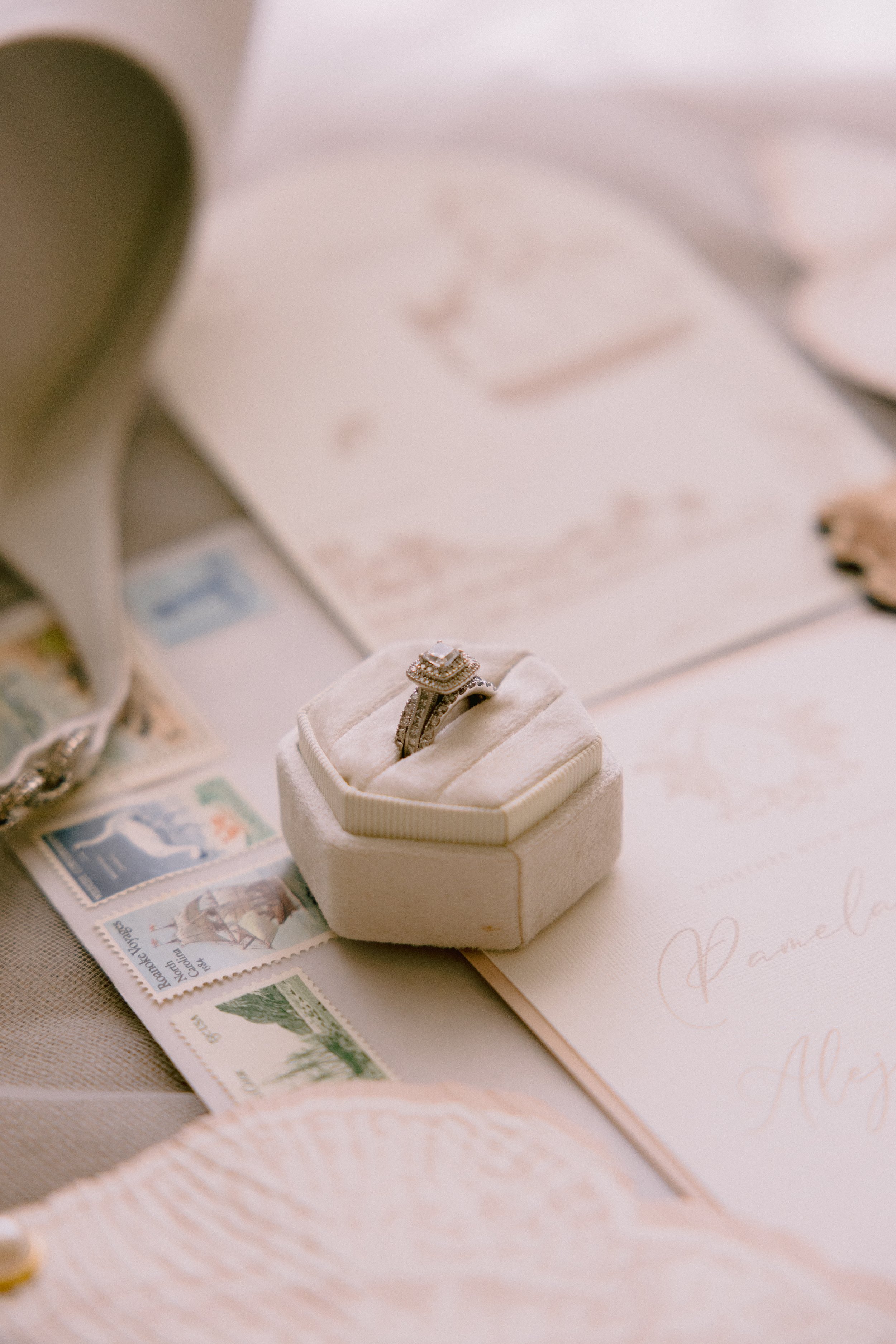A diamond engagement ring in a white velvet ring box, with wedding invitations and postage stamps nearby.