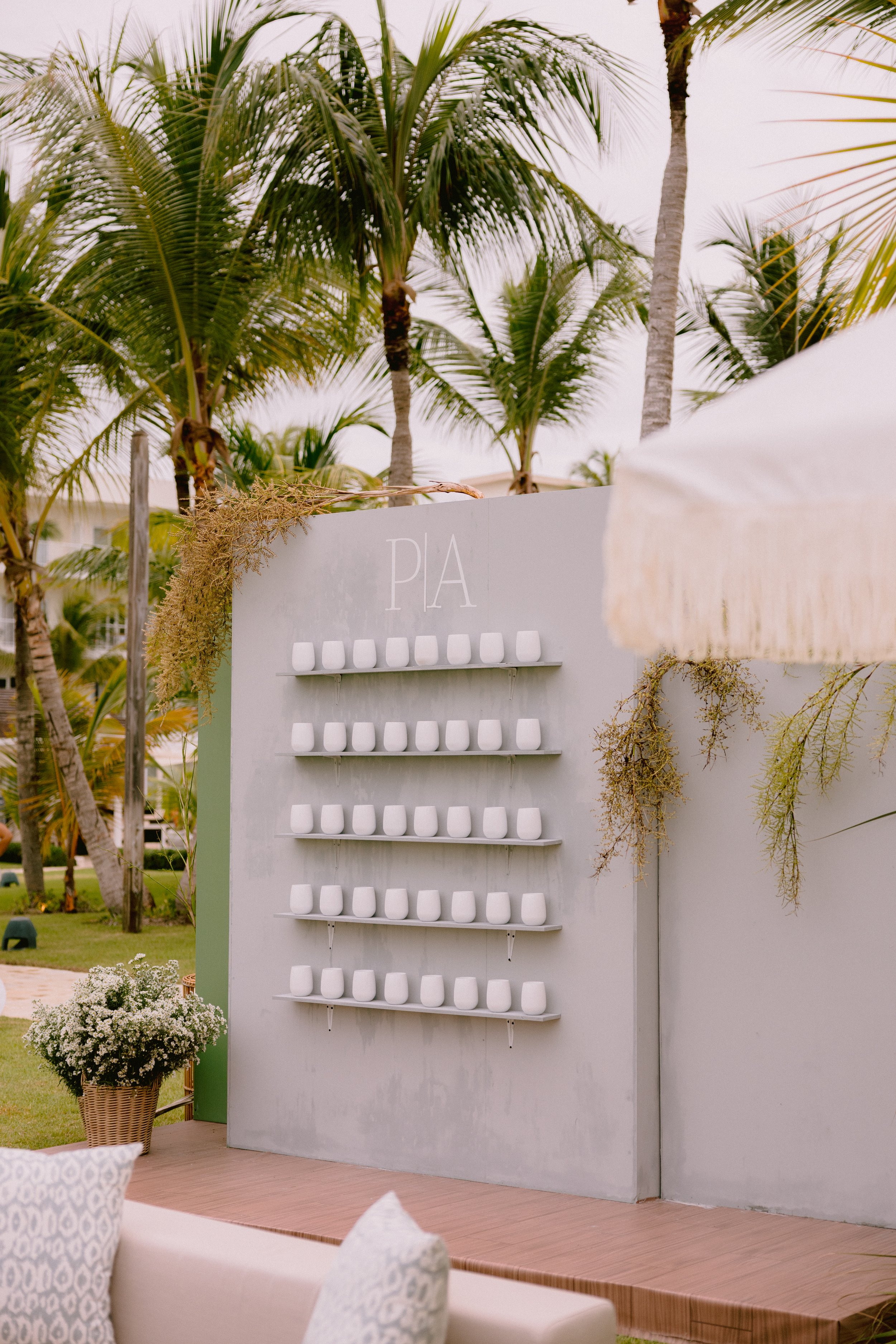 White wall with the word 'PIA' and five shelves holding white cups, outdoors with palm trees and a parasol nearby.
