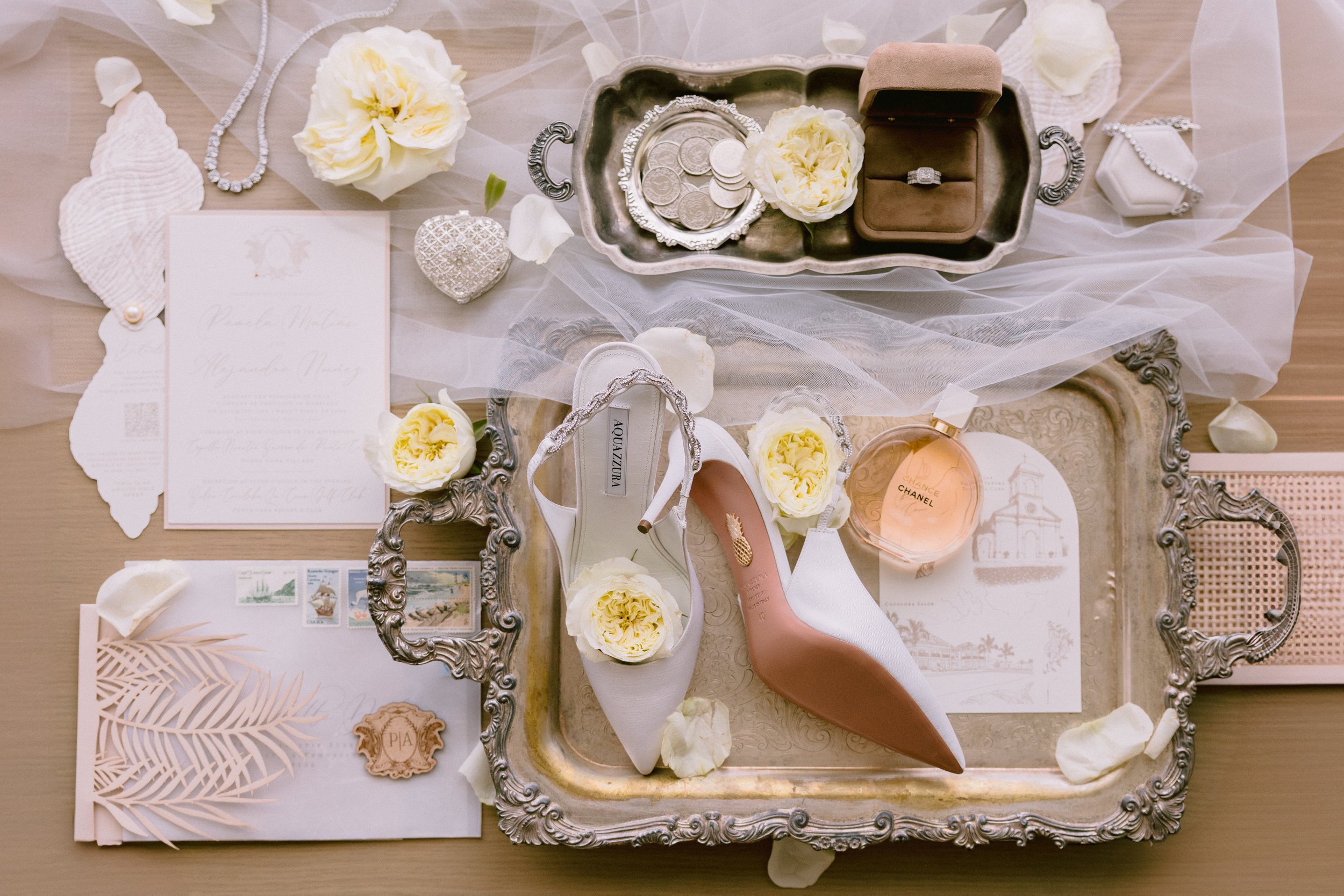 Wedding accessories including white and blush high-heeled shoes, a Chanel perfume bottle, pearl-embellished jewelry, a silver tray, a ring in a box, flowers, a wedding invitation, and decorative items on a silver tray and wooden surface.
