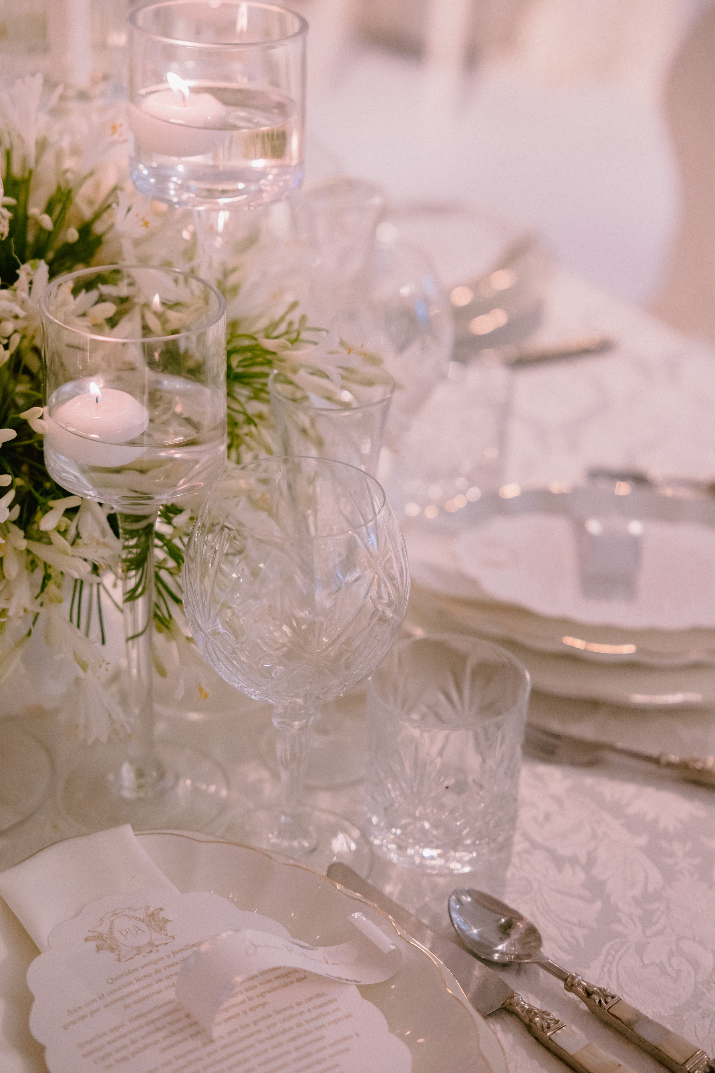 Elegant table setting with white floral centerpiece, candles, crystal glasses, silverware, and a printed menu on a white tablecloth.