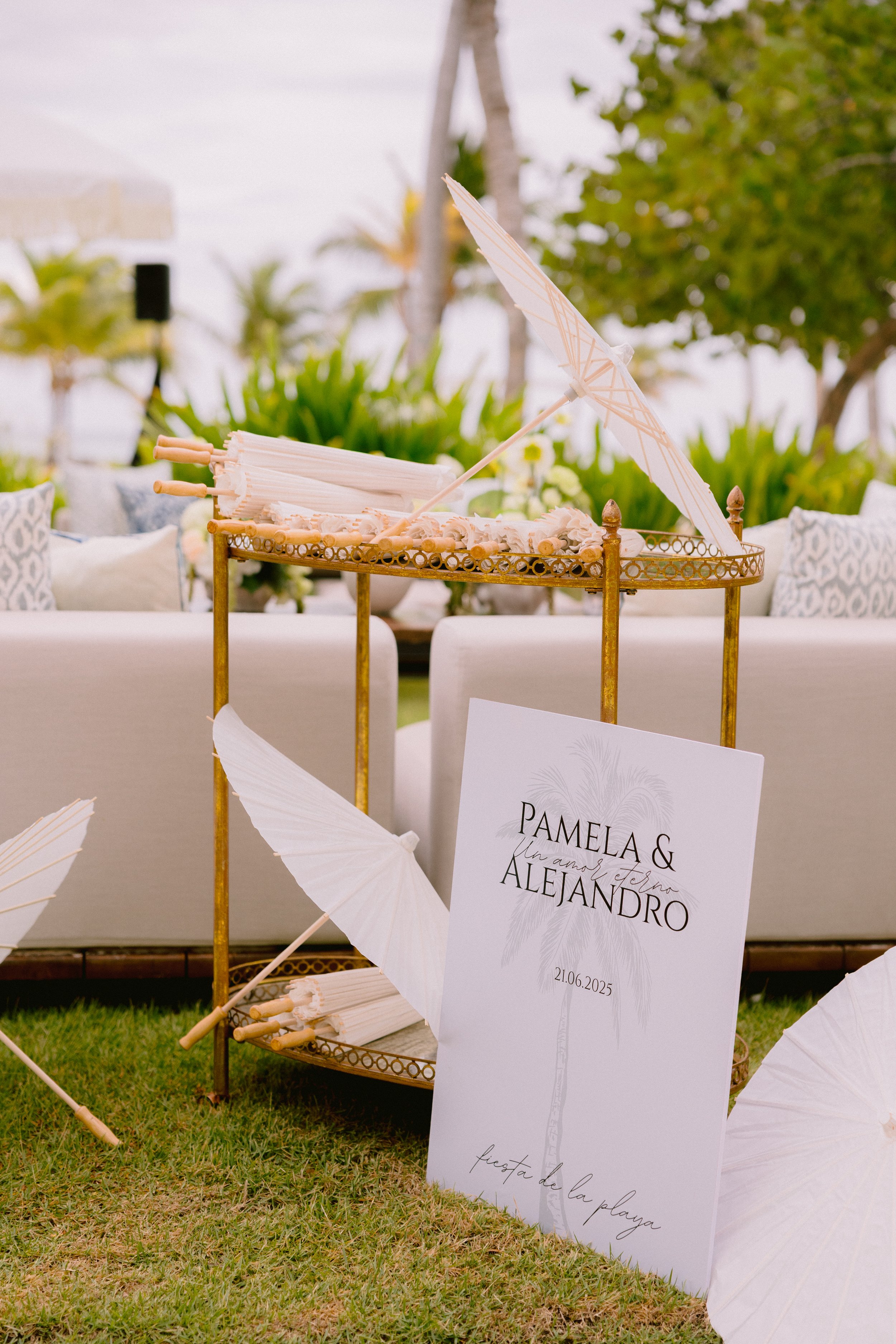 Wedding reception decor featuring a small golden cart, decorated with paper umbrellas and fans, with a sign that reads 'Pamela & Alejandro, 21.06.2025, fiesta de la playa' on a grassy area, with outdoor seating and greenery in the background.