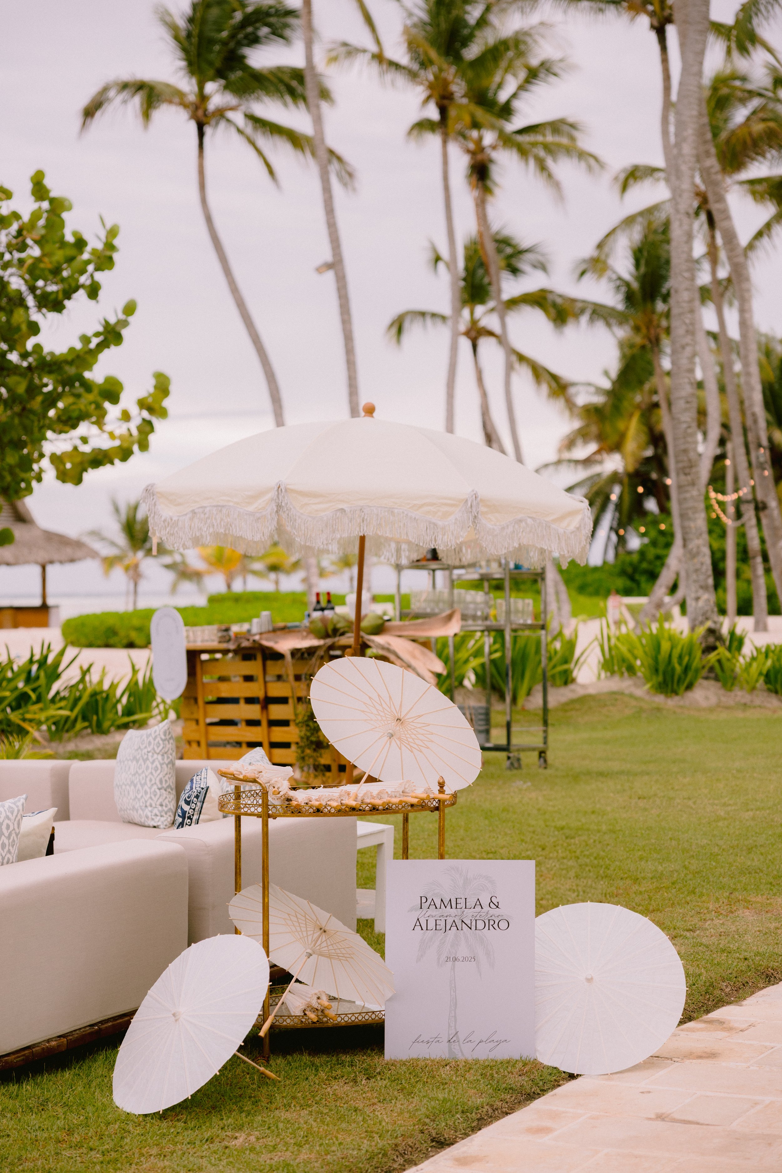 Wedding reception setup on a beach with white umbrellas, a sign with names Pamela and Alejandro, and tropical palm trees in the background.