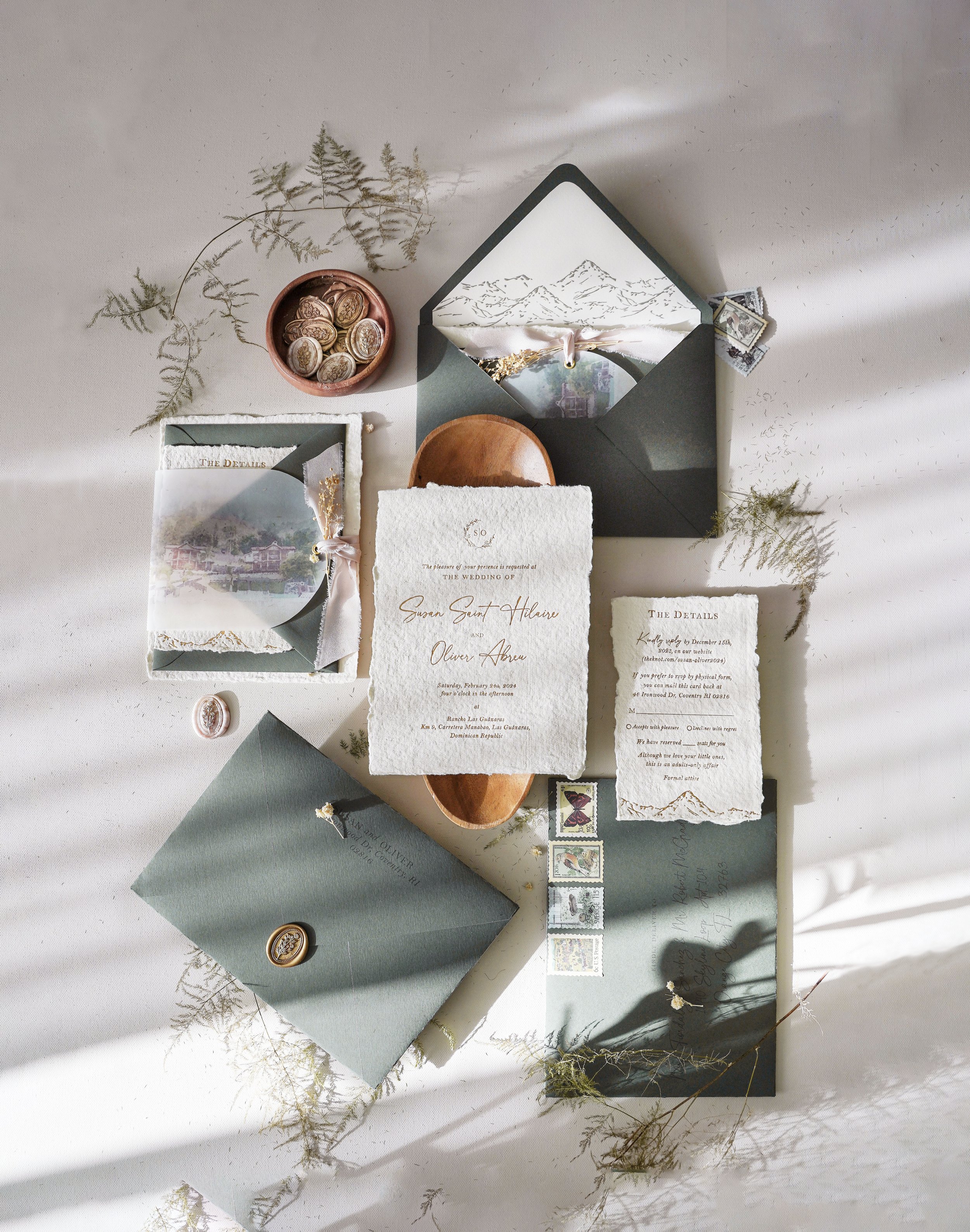 Wedding invitation suite with textured papers, envelopes, stamps, and dried botanical accents arranged on a white surface.