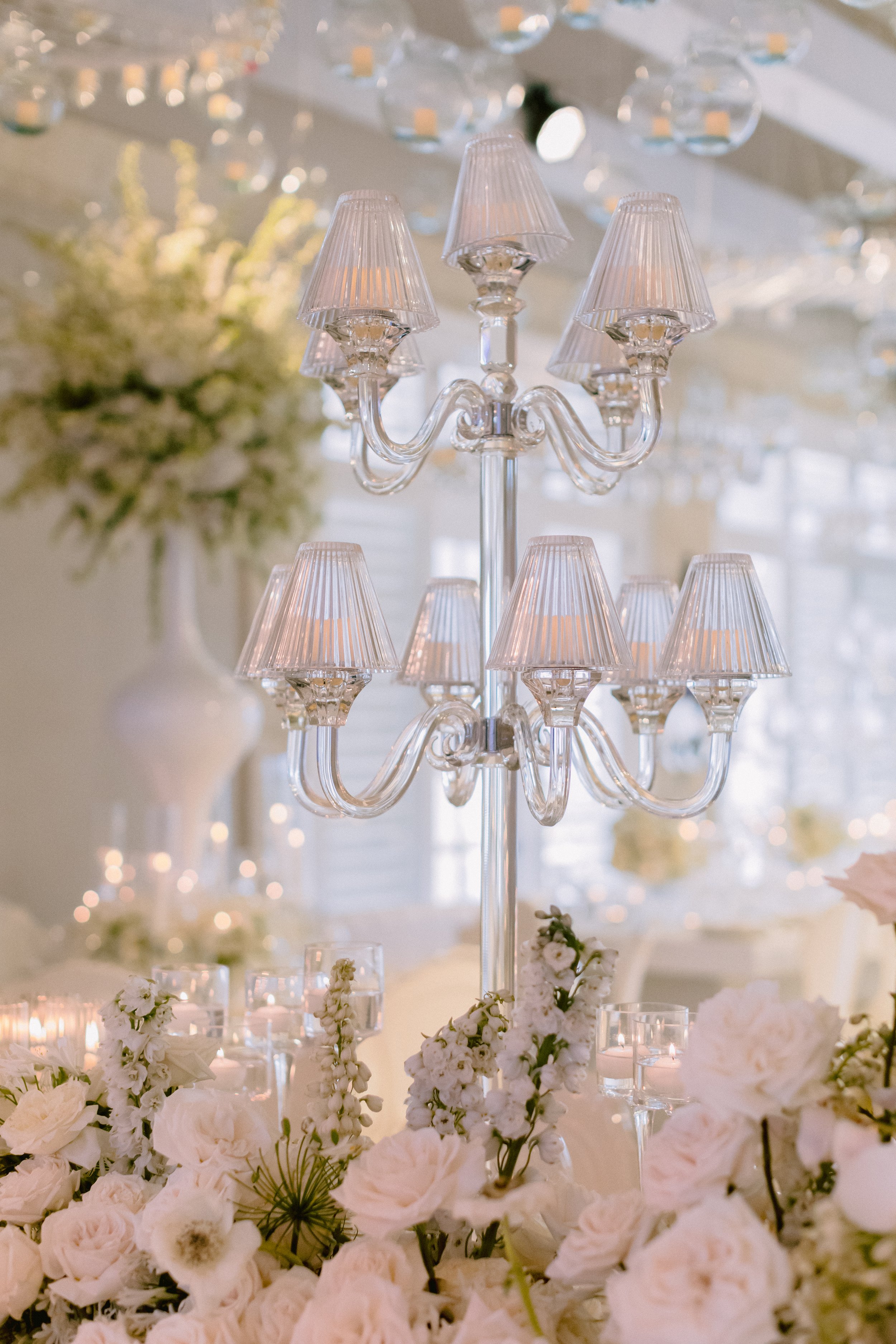 Elegant chandelier with multiple arms and small shades, surrounded by white flowers and candles in a decorated event space.