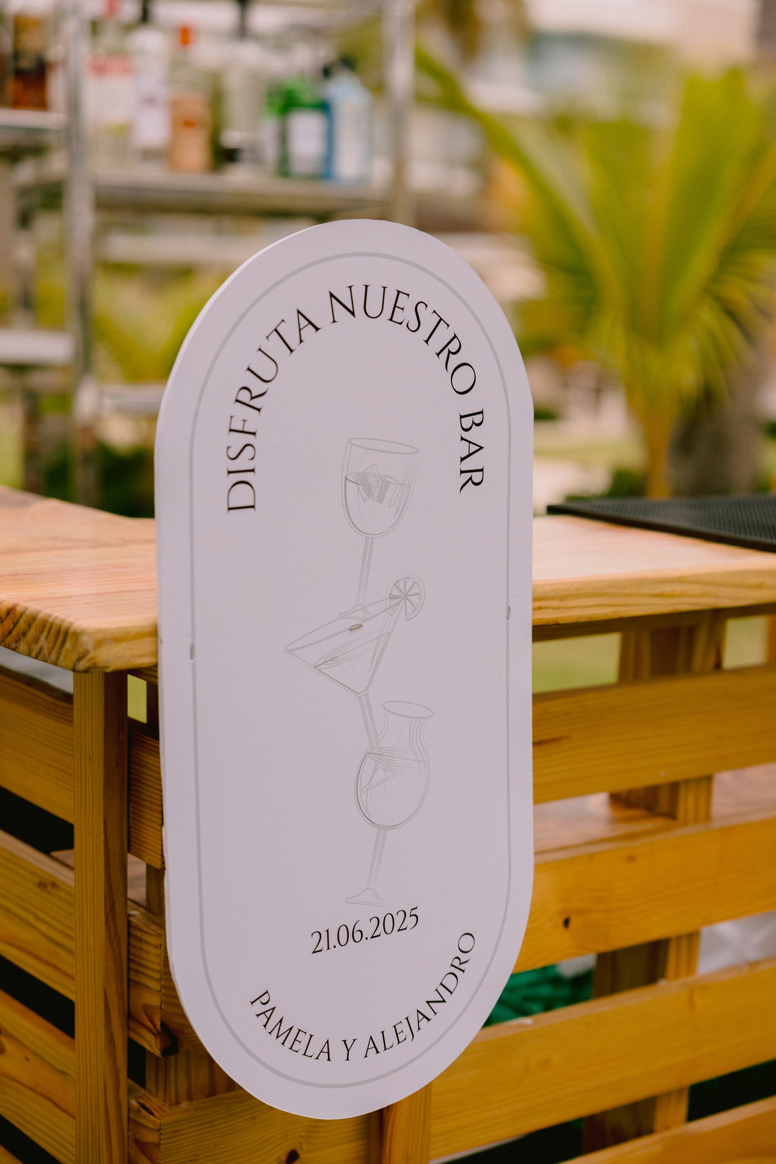 Wedding bar sign with illustrated cocktail glasses, the date 21.06.2025, and the names Pamela and Alejandro, displayed on a wooden bar counter with a blurred outdoor background.