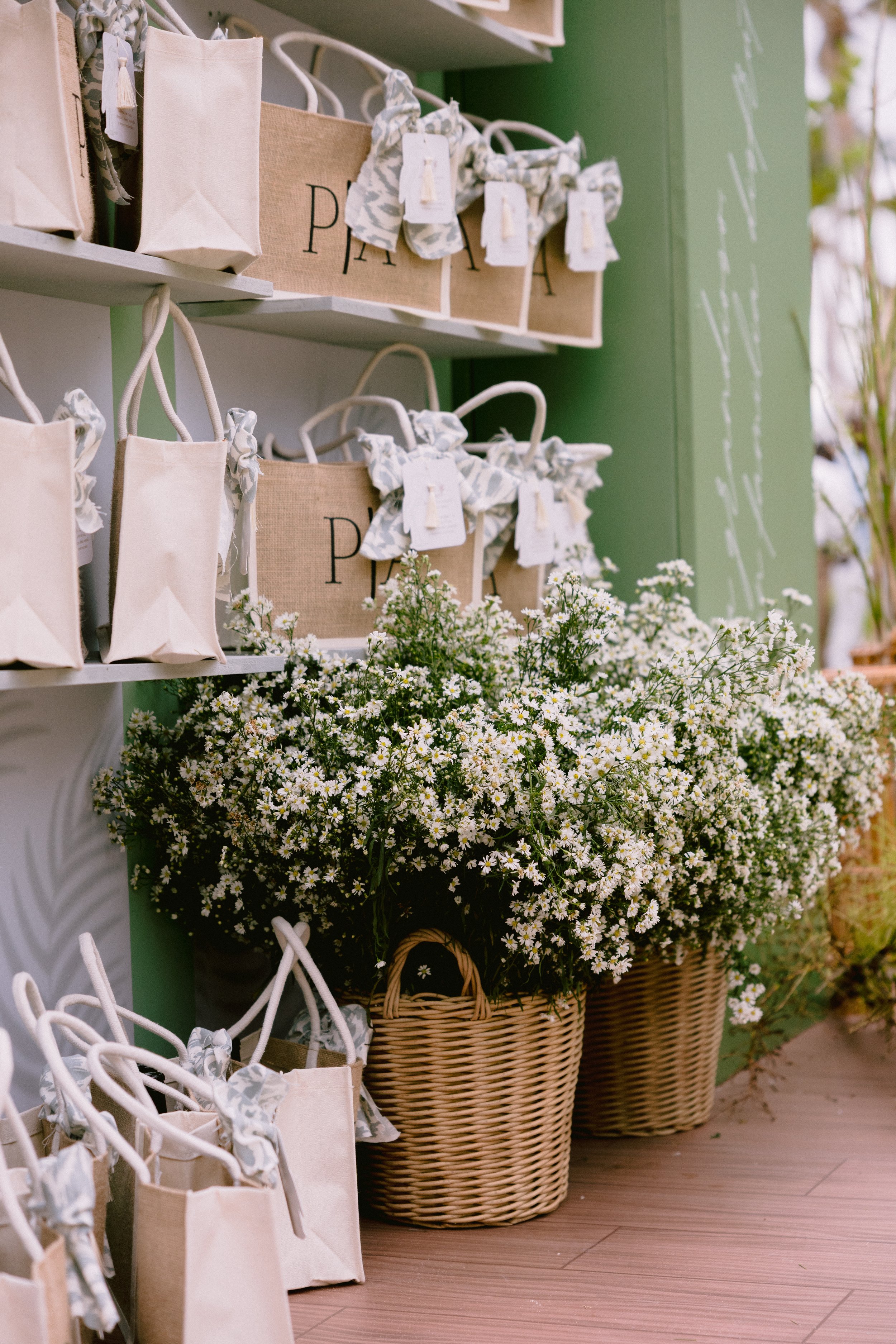 Display of white and beige gift bags with floral bows, placed on shelves, and large pots filled with white daisies, all against a green wall at a store.