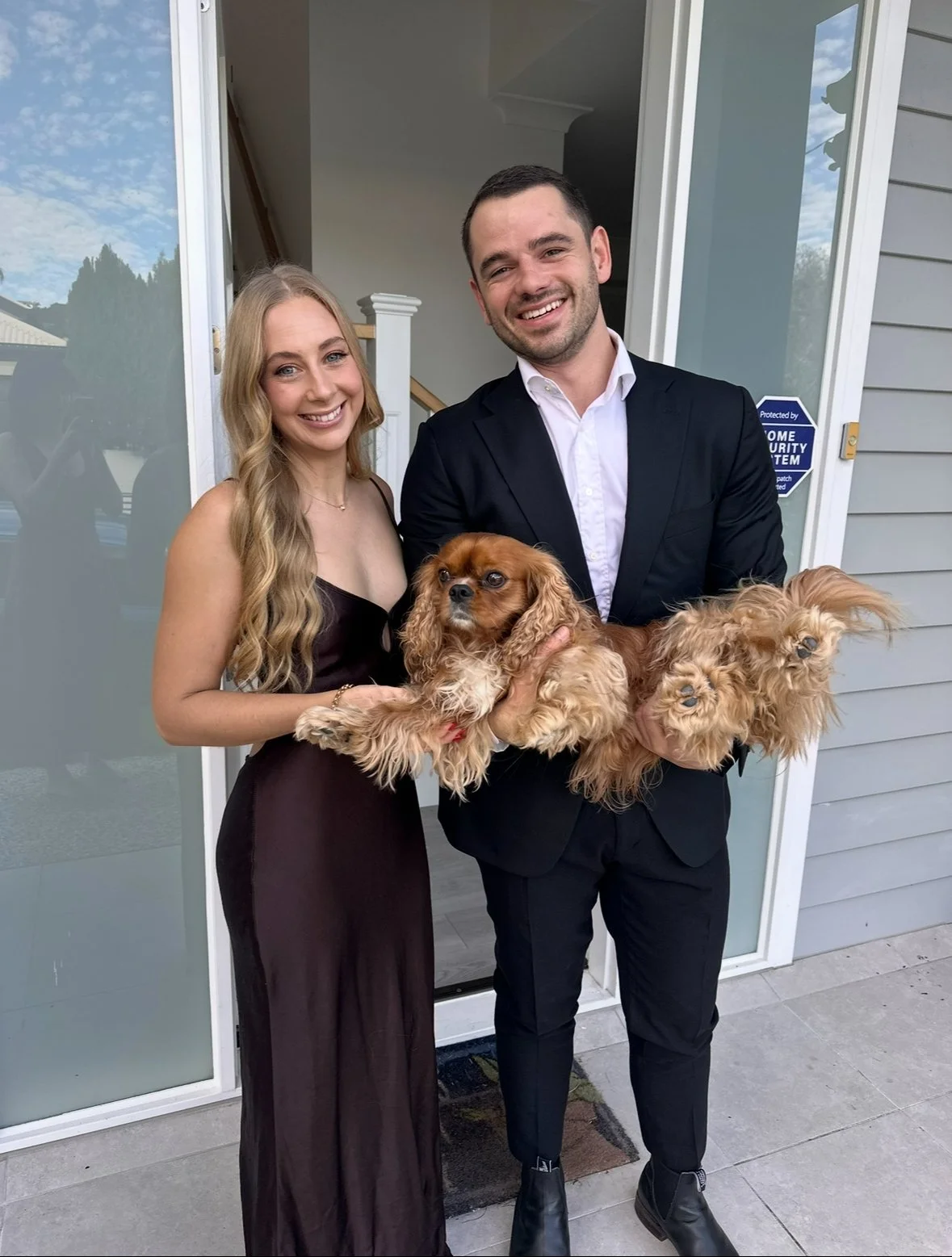 A smiling man and woman standing at a house doorway, holding two small brown dogs.
