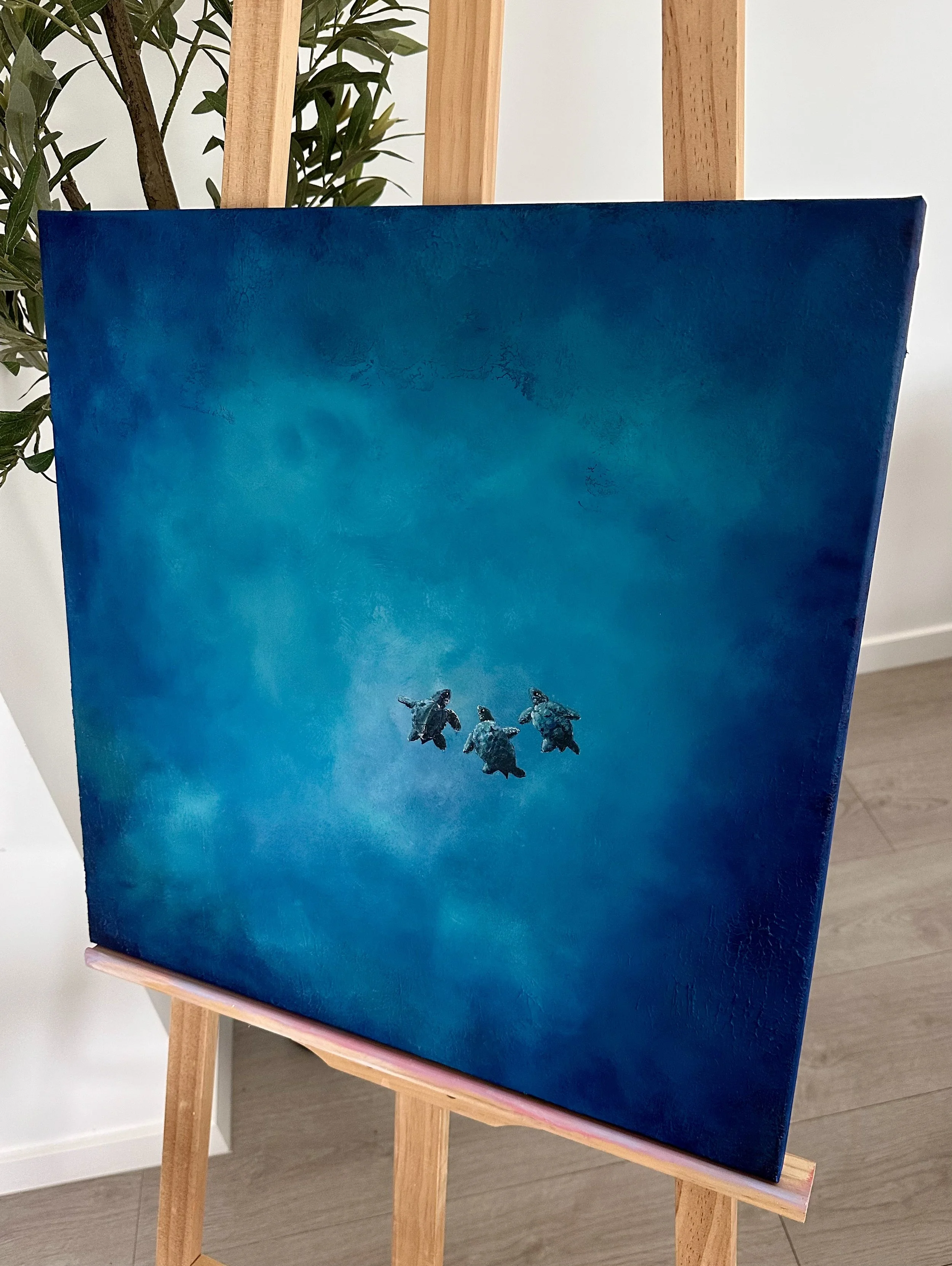 A painting on a canvas depicts four turtles swimming underwater with a blue background. The canvas is displayed on a wooden easel in a room with white walls and wooden flooring, with some greenery visible behind the easel.