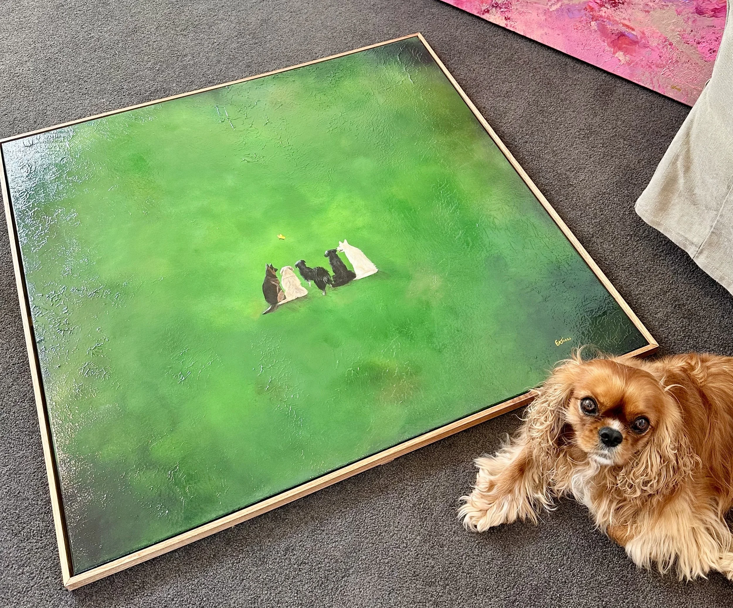 A painting of five dogs sitting on a green field with trees and a yellow butterfly, placed on a dark carpeted floor. Part of a pink painting and a dog with long ears are also visible.