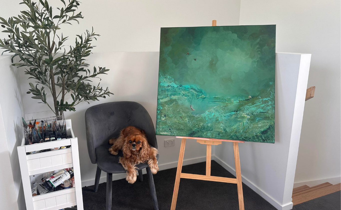 A dog sitting in a gray chair next to a white art supply storage cart with brushes and paint on it, a large green abstract painting on an easel, a potted plant, and a white wall.