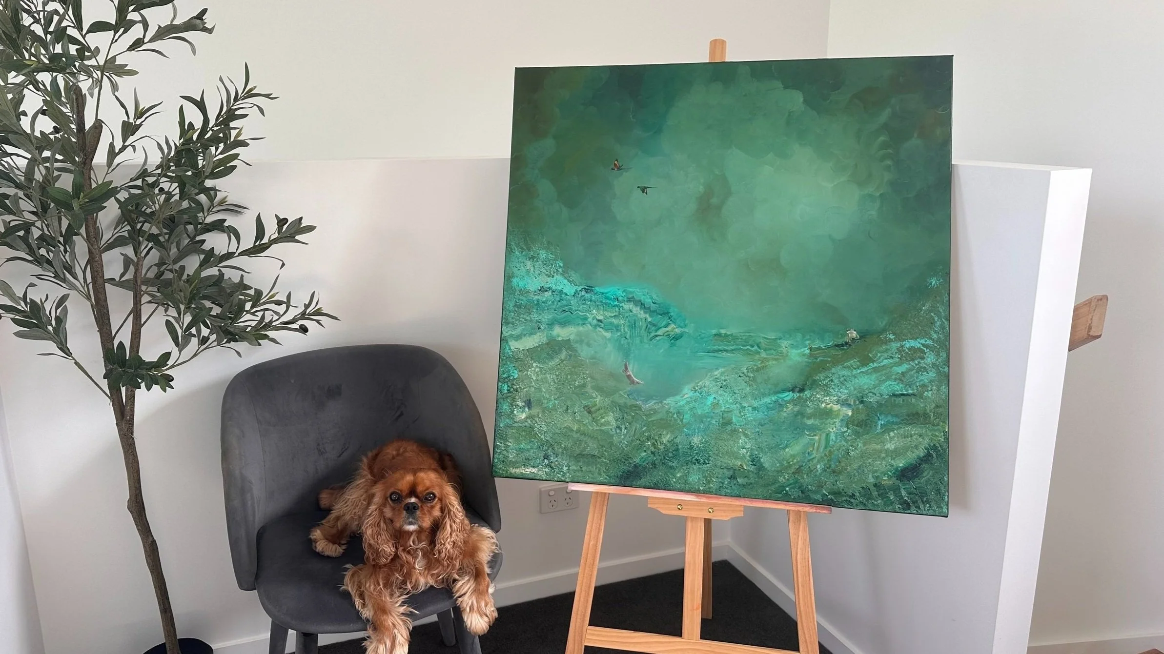 A dog sitting on a gray chair next to a green plant, with an abstract green painting on an easel behind them in a room with white walls.