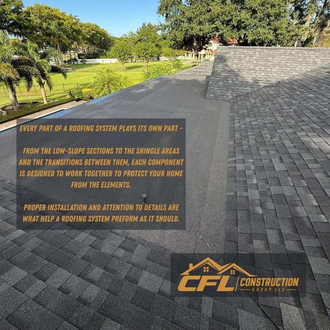 View of a rooftop with asphalt shingles, surrounded by lush green trees and a fenced backyard, under a clear blue sky.
