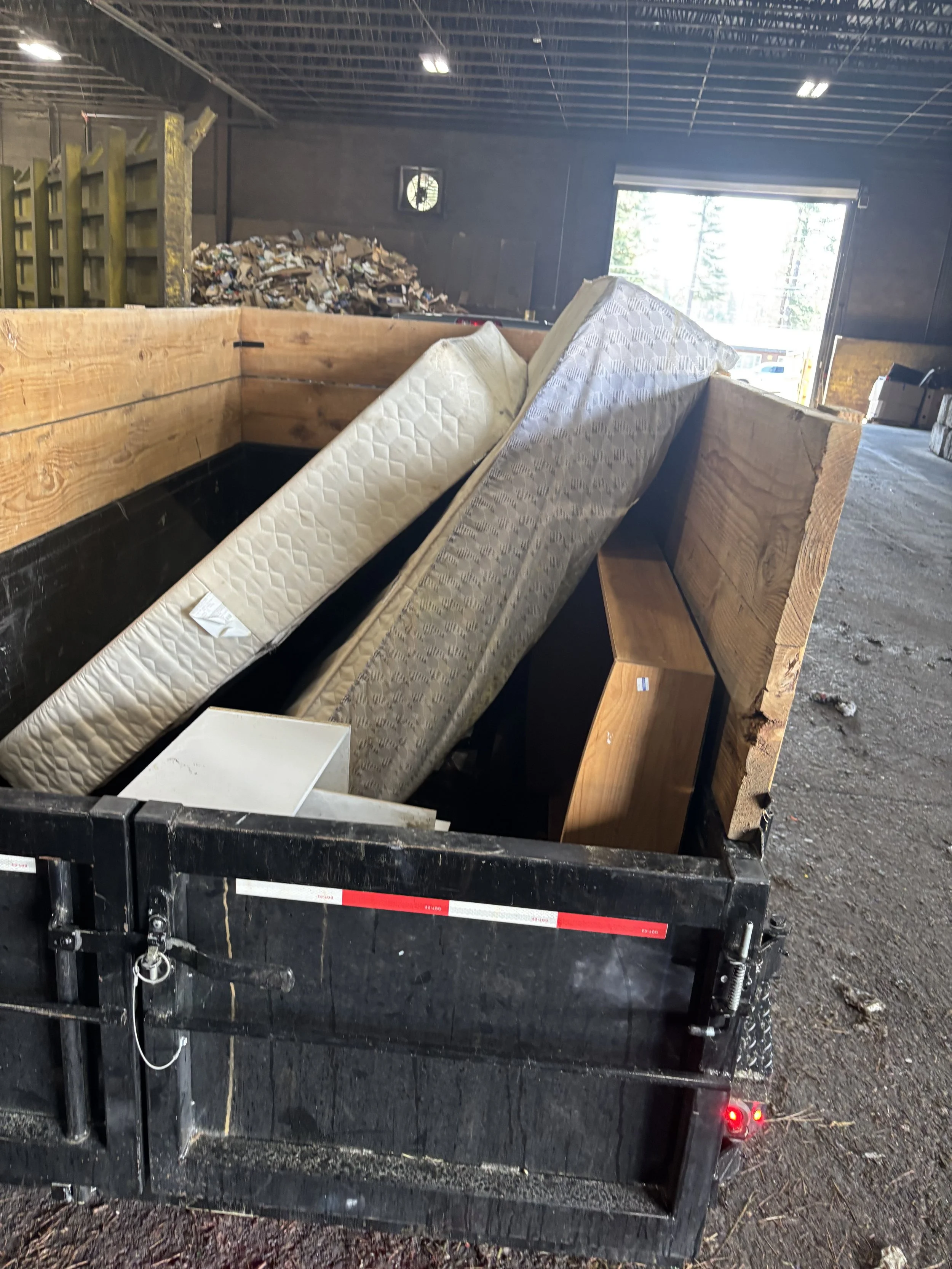 View of a trailer loaded with mattresses inside a warehouse with piles of trash and a clock on the wall.