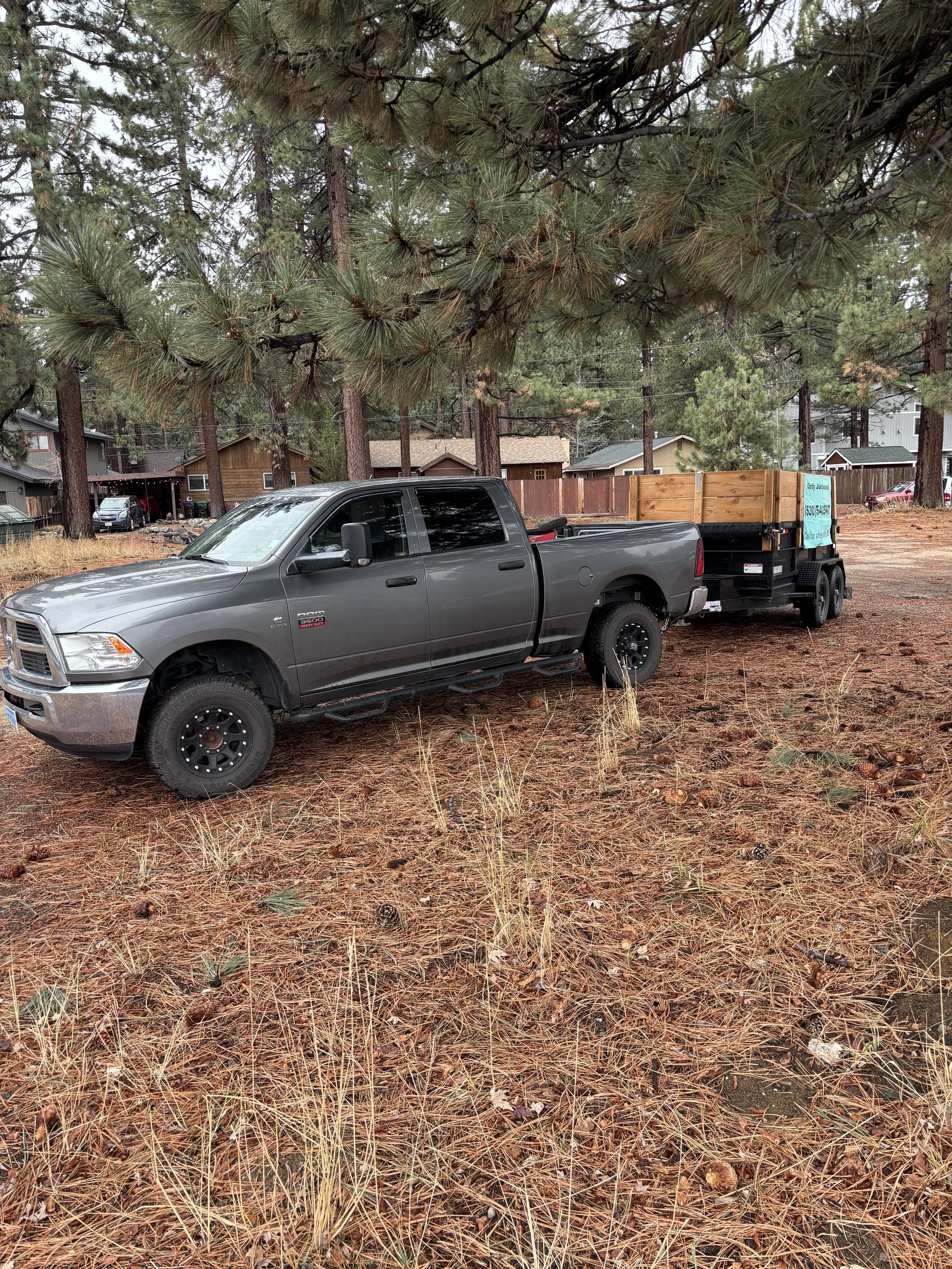 A gray pickup truck with a trailer parked on a grassy, pine-tree forested area with houses in the background.