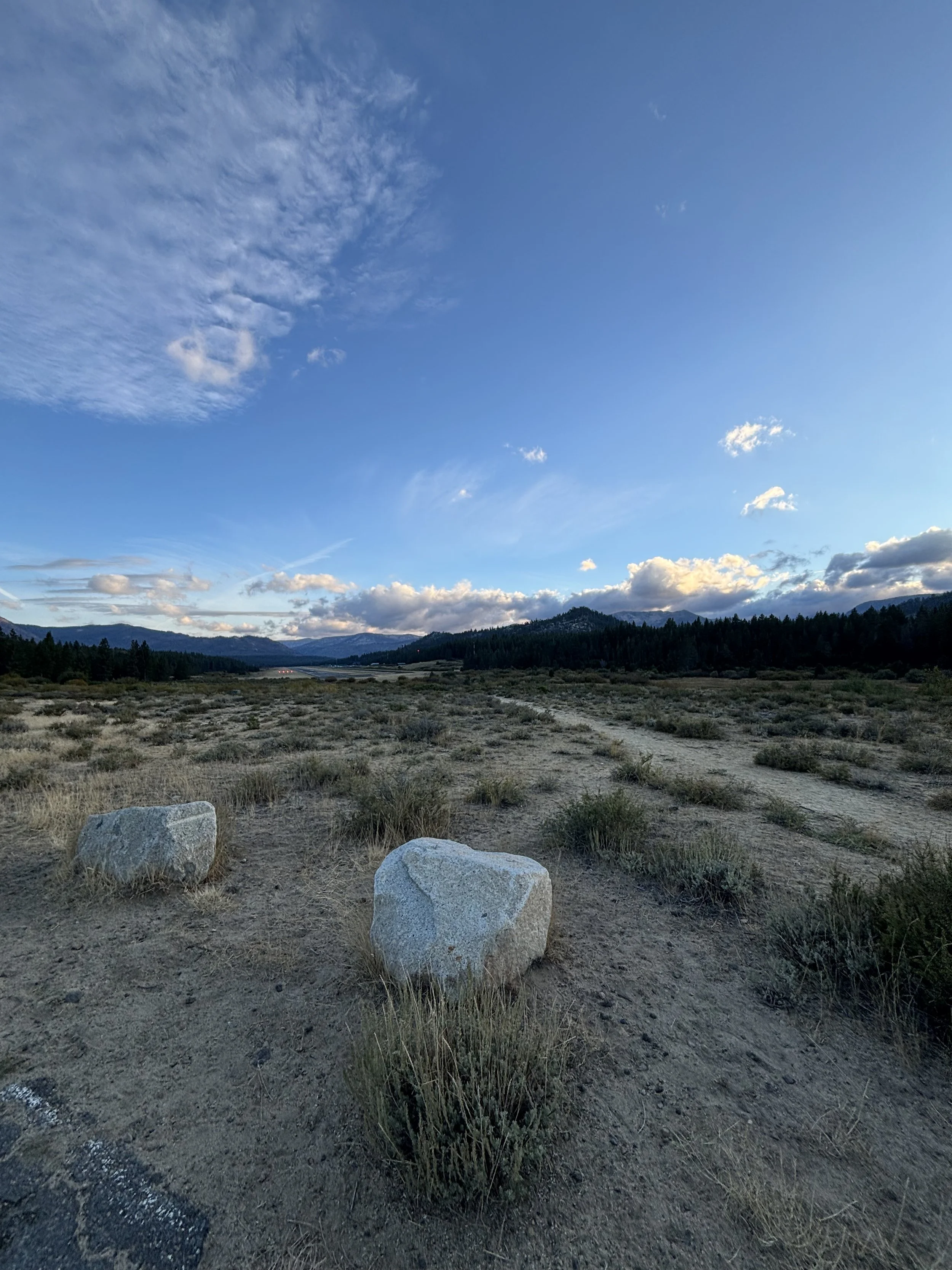 Open landscape with dirt road, scattered rocks, dry grass, distant mountains, and a sky with light clouds at dusk or dawn.