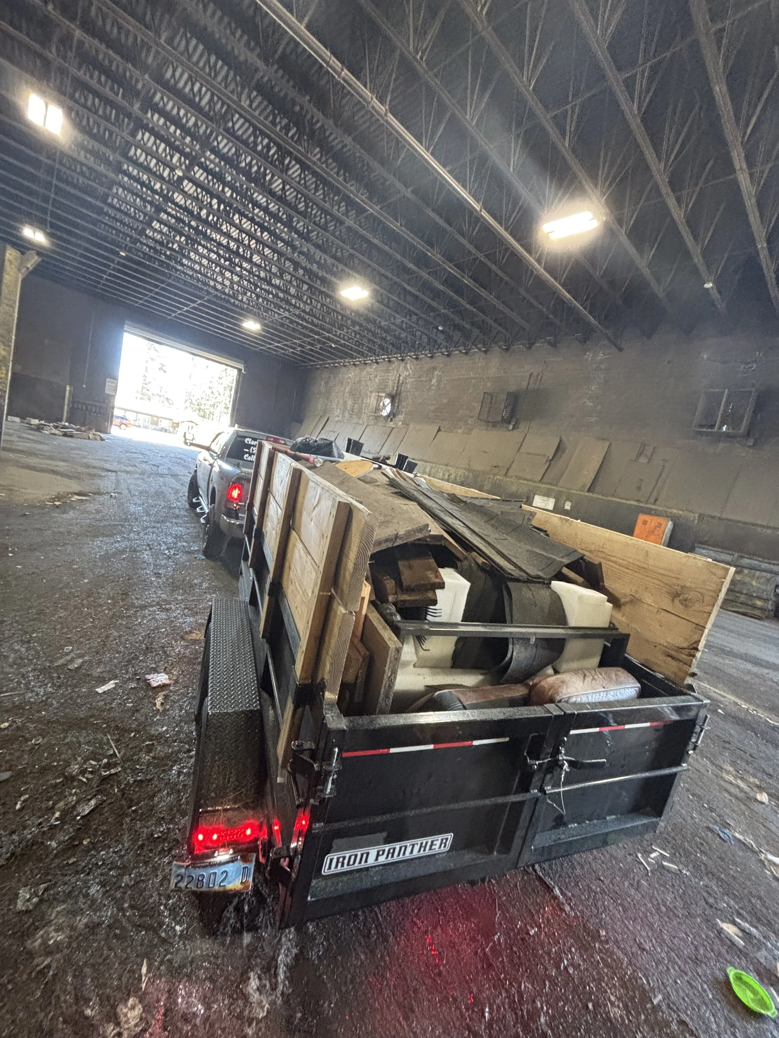 The image shows the interior of a large warehouse or industrial building with a high, curved metal ceiling. There is a small utility trailer filled with various wooden planks, some furniture, and other debris. The trailer has the words 'IRON PANTHER'