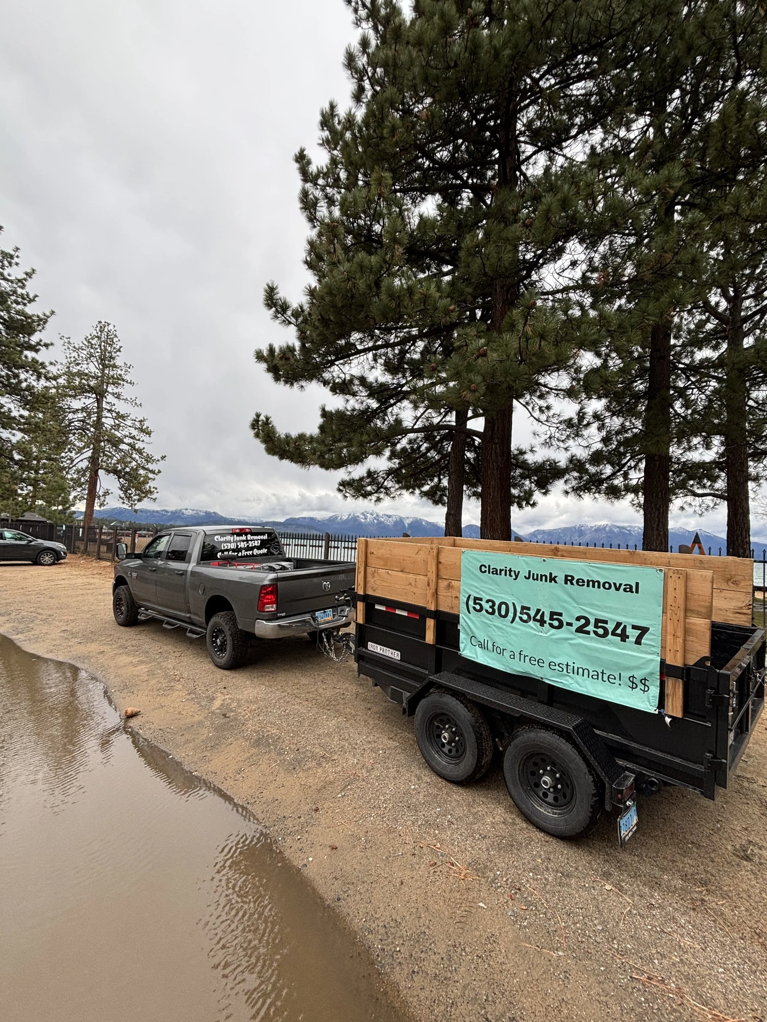 A truck trailer with a sign advertising Clarity Junk Removal, parked near a muddy, waterlogged area with trees and mountains in the background.