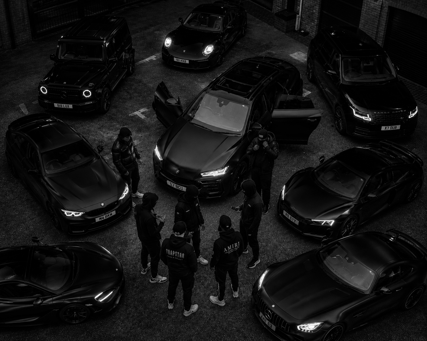 A group of people standing around a black Tesla Model S parked in a lot, surrounded by several other luxury cars including Range Rovers, Porsches, BMWs, and Audis, in a dark urban setting.