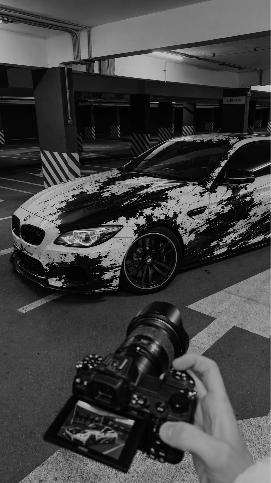 Black and white photo of a sports car with a splatter paint design in an underground parking garage, a person holding a camera in the foreground takes a picture of the car.