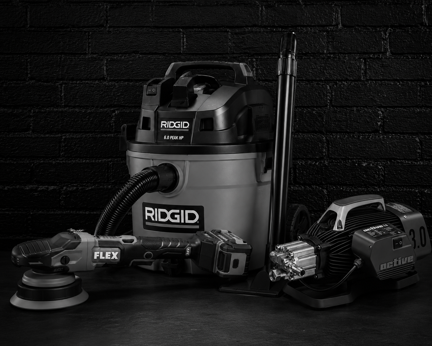 Black and gray Ridgid wet-dry vacuum, a cordless electric polisher, and an air compressor on a black wooden surface with a black brick wall background.