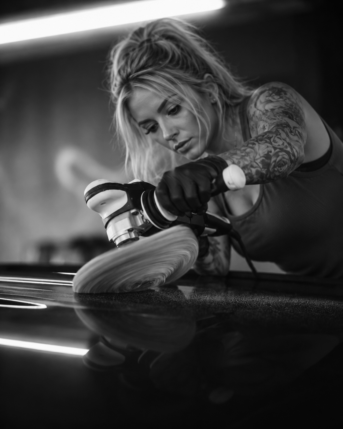 A woman with tattoos on her arms polishing a car with a polishing machine.