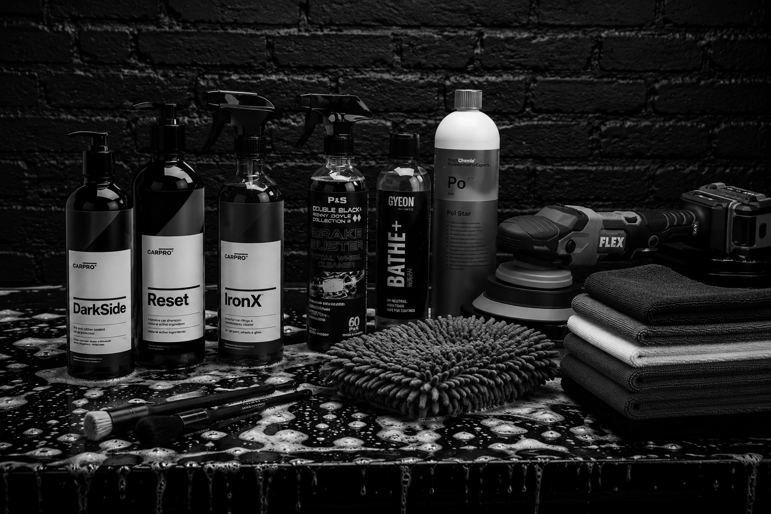 Car detailing supplies including bottles labeled DarkSide, Reset, IronX, P&S Brake Buster, GYEON Bathe+; a spray gun; a polishing machine; rolled towels; brushes; and sponges on a wet surface against a brick wall.