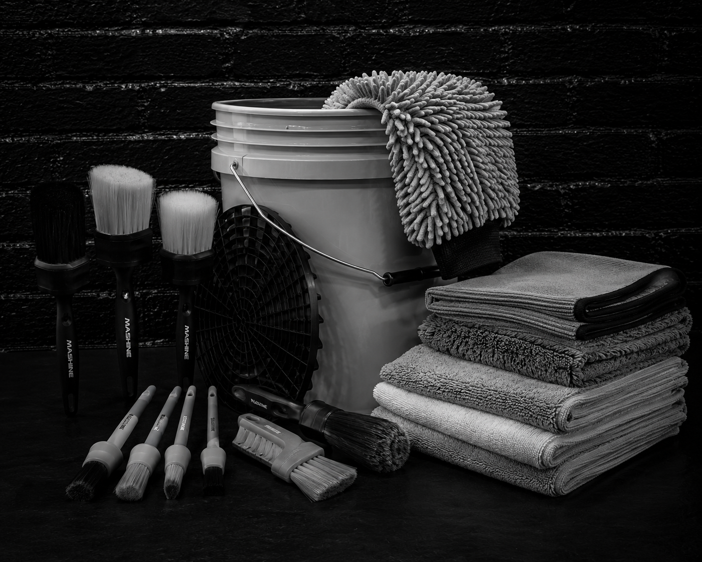 Collection of cleaning tools including brushes, a mop, a fan, stacked towels, and a bucket against a dark brick wall.