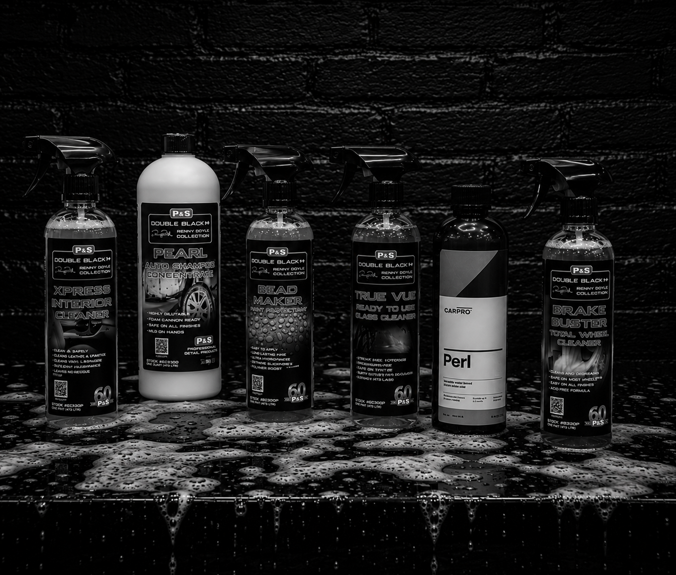 Collection of car cleaning products including interior cleaner, auto shampoo, bead maker, glass cleaner, perceived to be a polishing product, and brake cleaner, all on a wet surface with soapy water and foam.