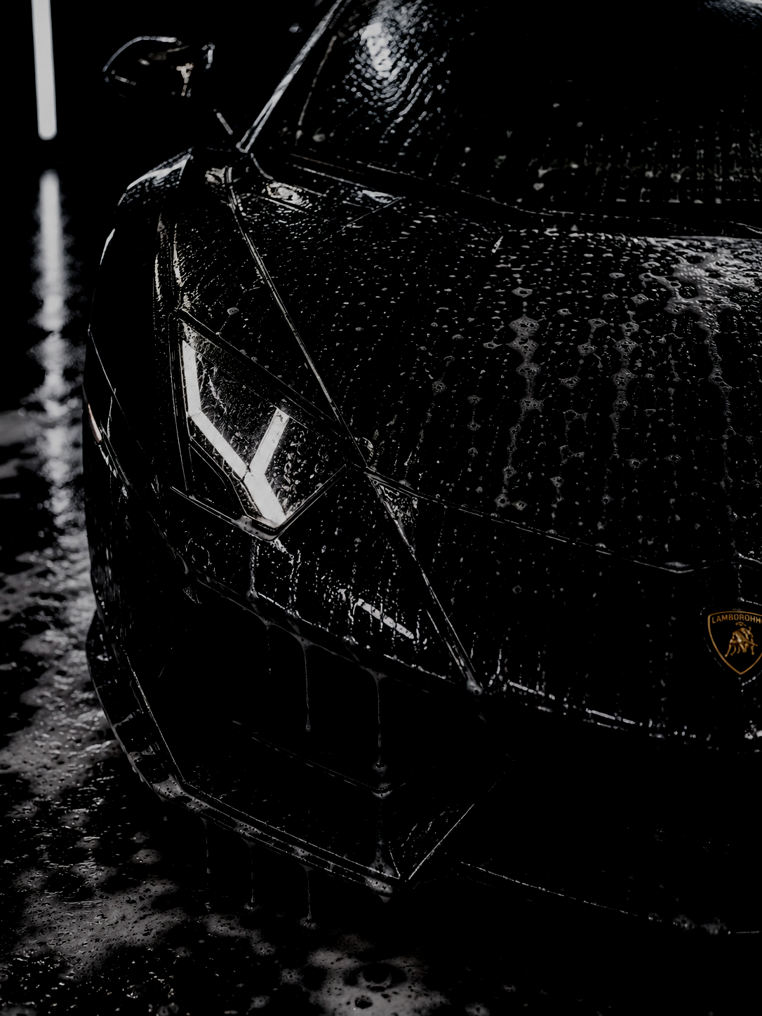 Close-up of a black Lamborghini luxury sports car covered in water droplets, with a soap bubble-laden surface, featuring a Lamborghini badge on the front. The car is wet from washing or rain, with dramatic lighting highlighting its sleek, angular design.