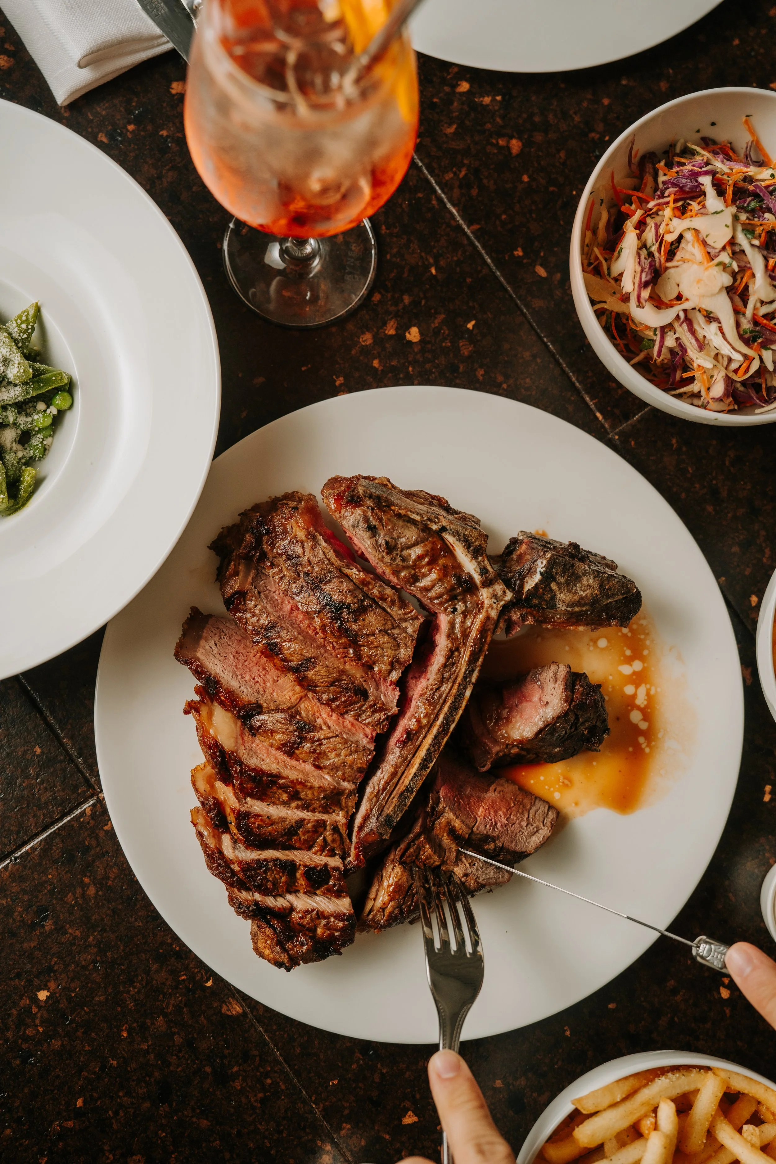 A dinner table with a roasted steak, a bowl of coleslaw, a bowl of pasta, and a glass of iced tea.