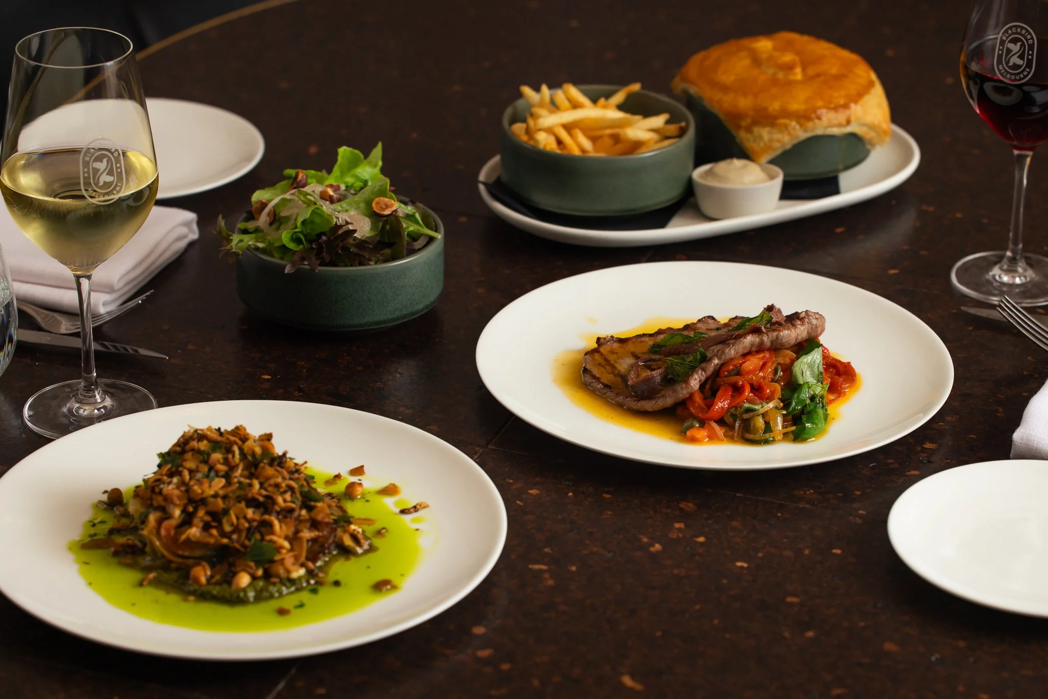 A dining table with plates of food, glasses of white and red wine, a green salad, French fries, a meat dish with vegetables, and a bread pudding dessert.