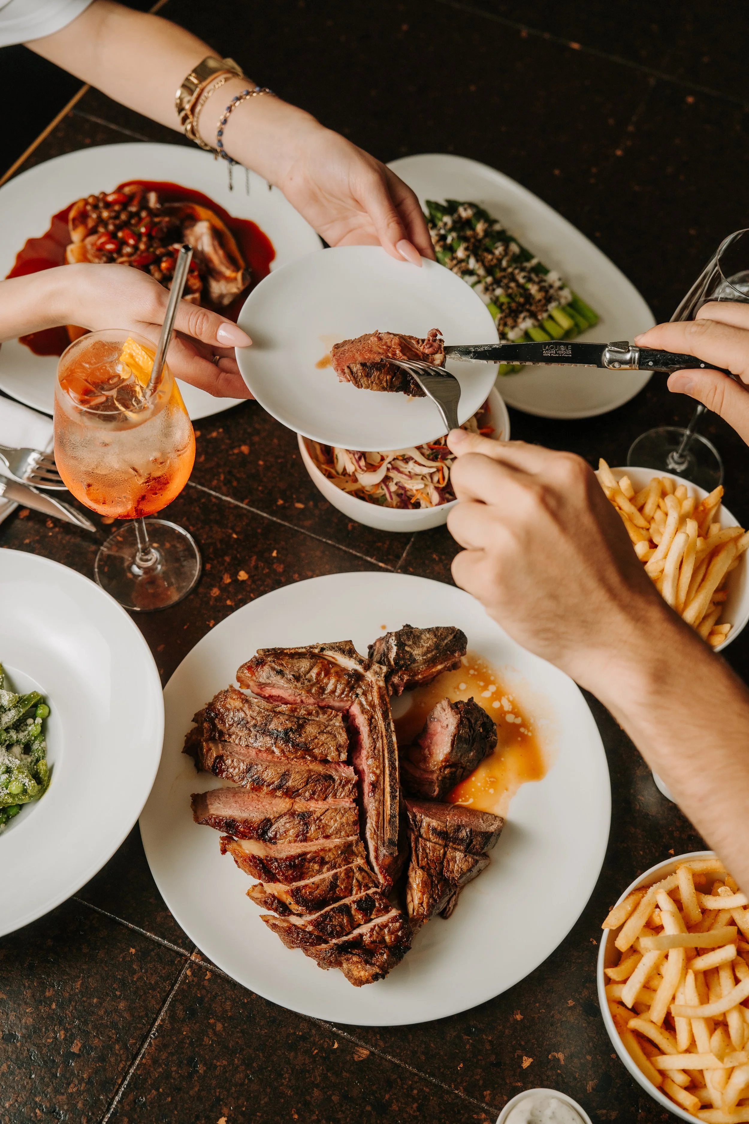People sharing a meal with grilled steak, salad, french fries, and drinks.