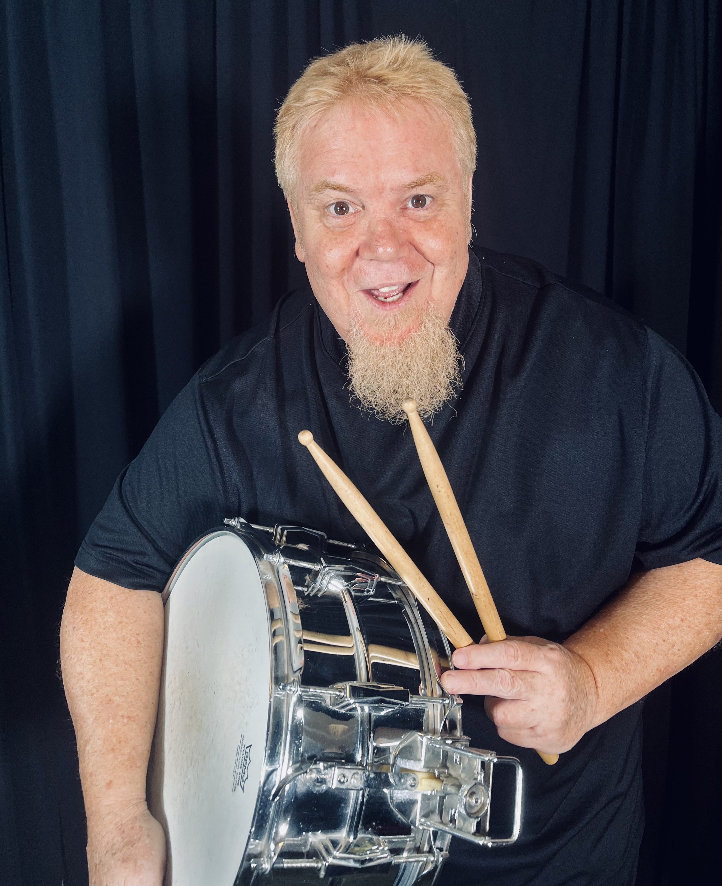 A man with blond hair and a goatee, wearing a black shirt, holding a snare drum and drumsticks, smiling with a dark background.