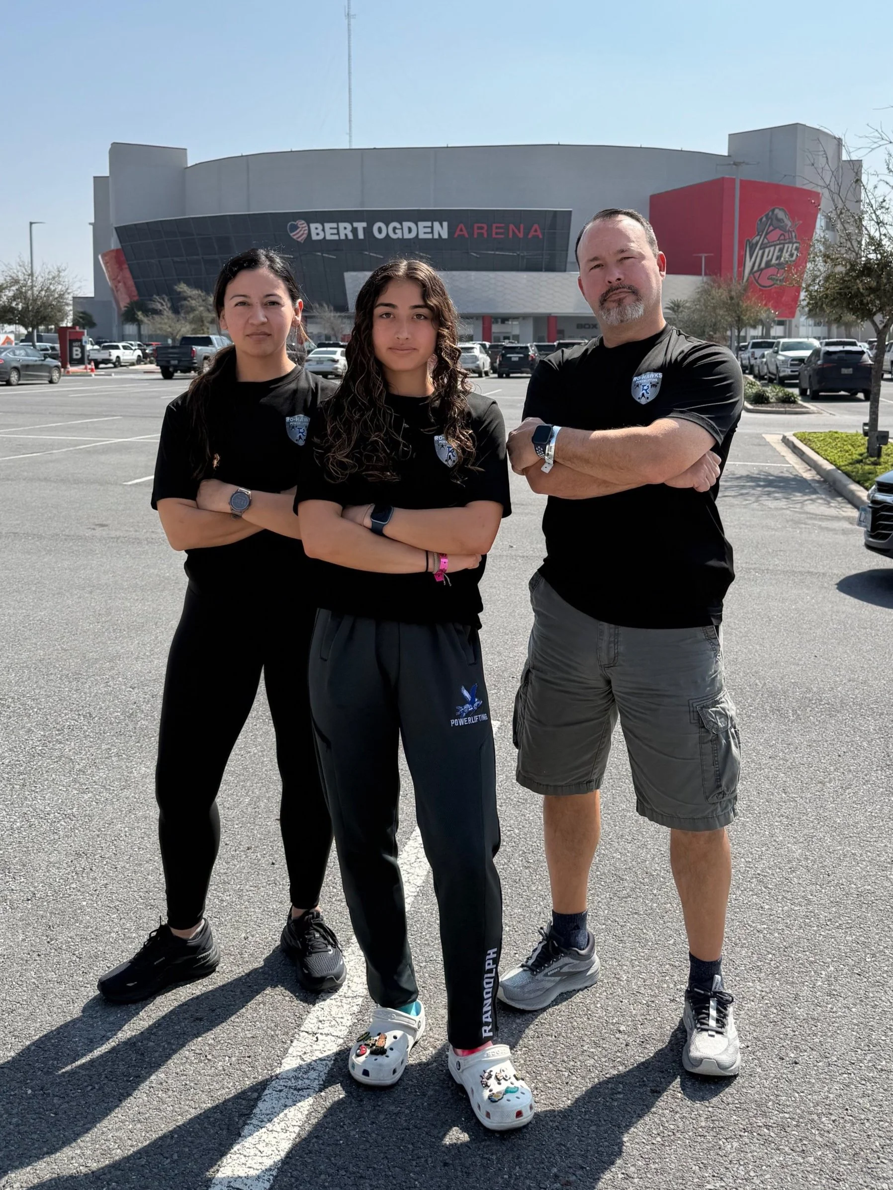 Three people standing with arms crossed in front of Bert Ogden Arena, wearing black shirts with team logo, in a parking lot on a sunny day.