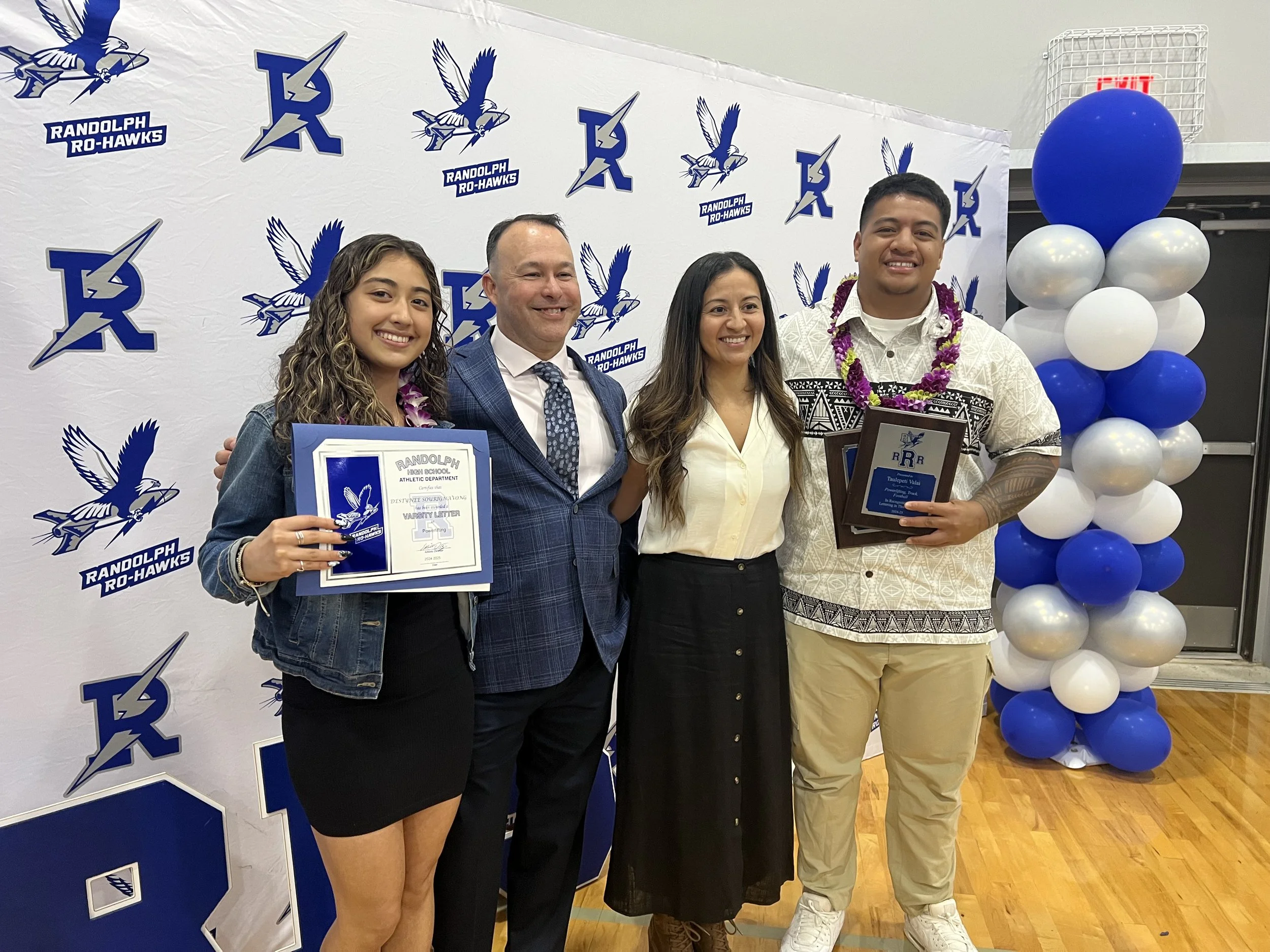 Group of four people standing together at an award ceremony, in front of a backdrop with the Randolph Ro-Hawks logo, some holding certificates, and there are balloons on the right side.