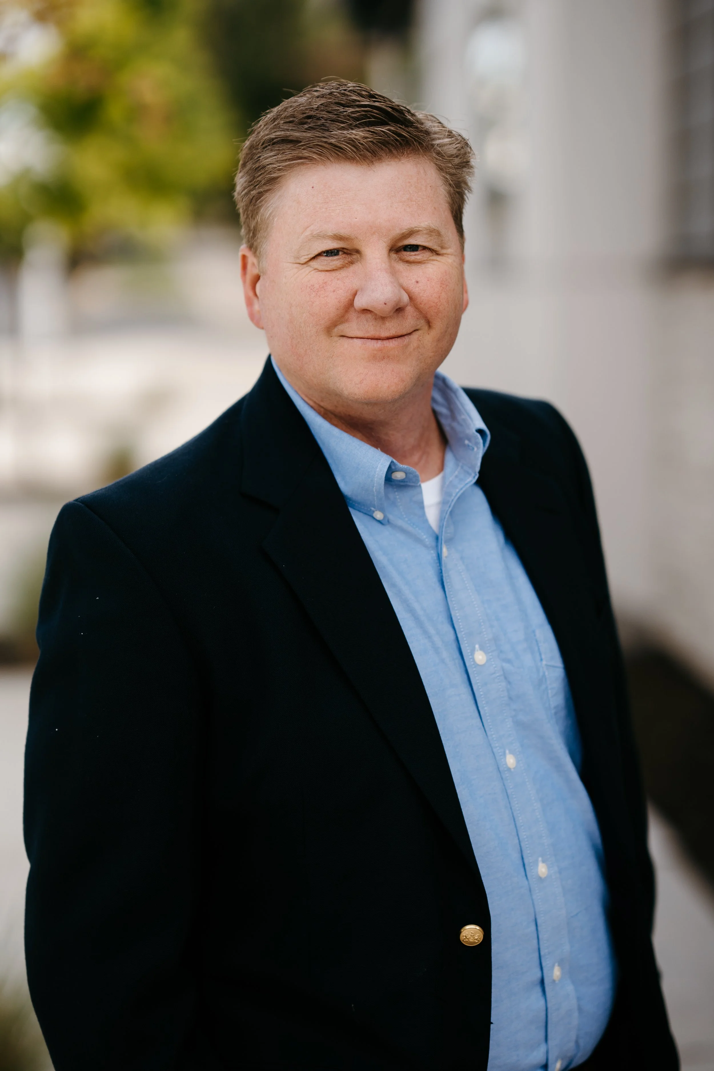 Portrait of a smiling man with short brown hair, dressed in a dark blazer and light blue shirt, standing outdoors.